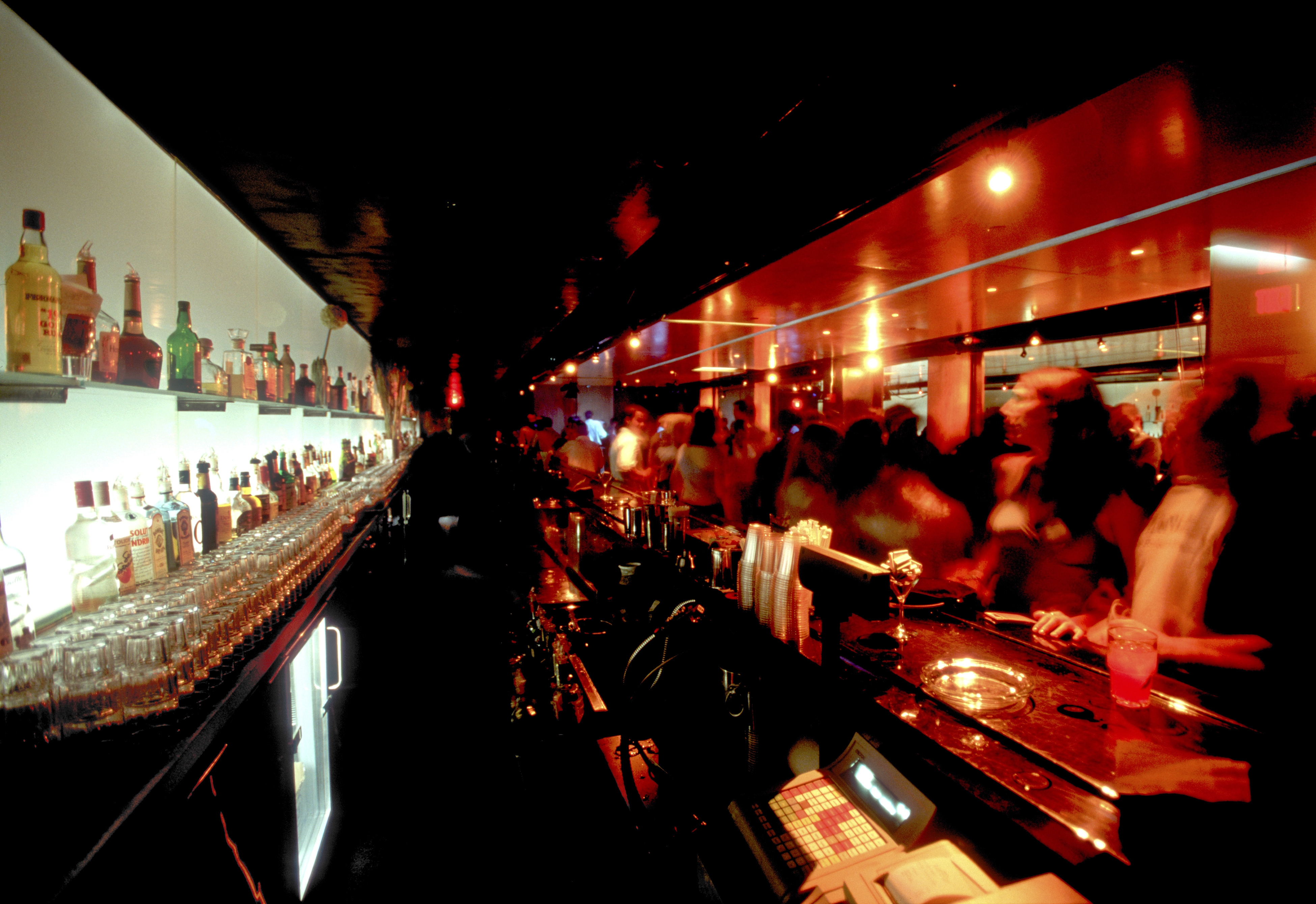 A busy bar with people socializing and various bottles of alcohol displayed behind the counter. The scene is lively and crowded