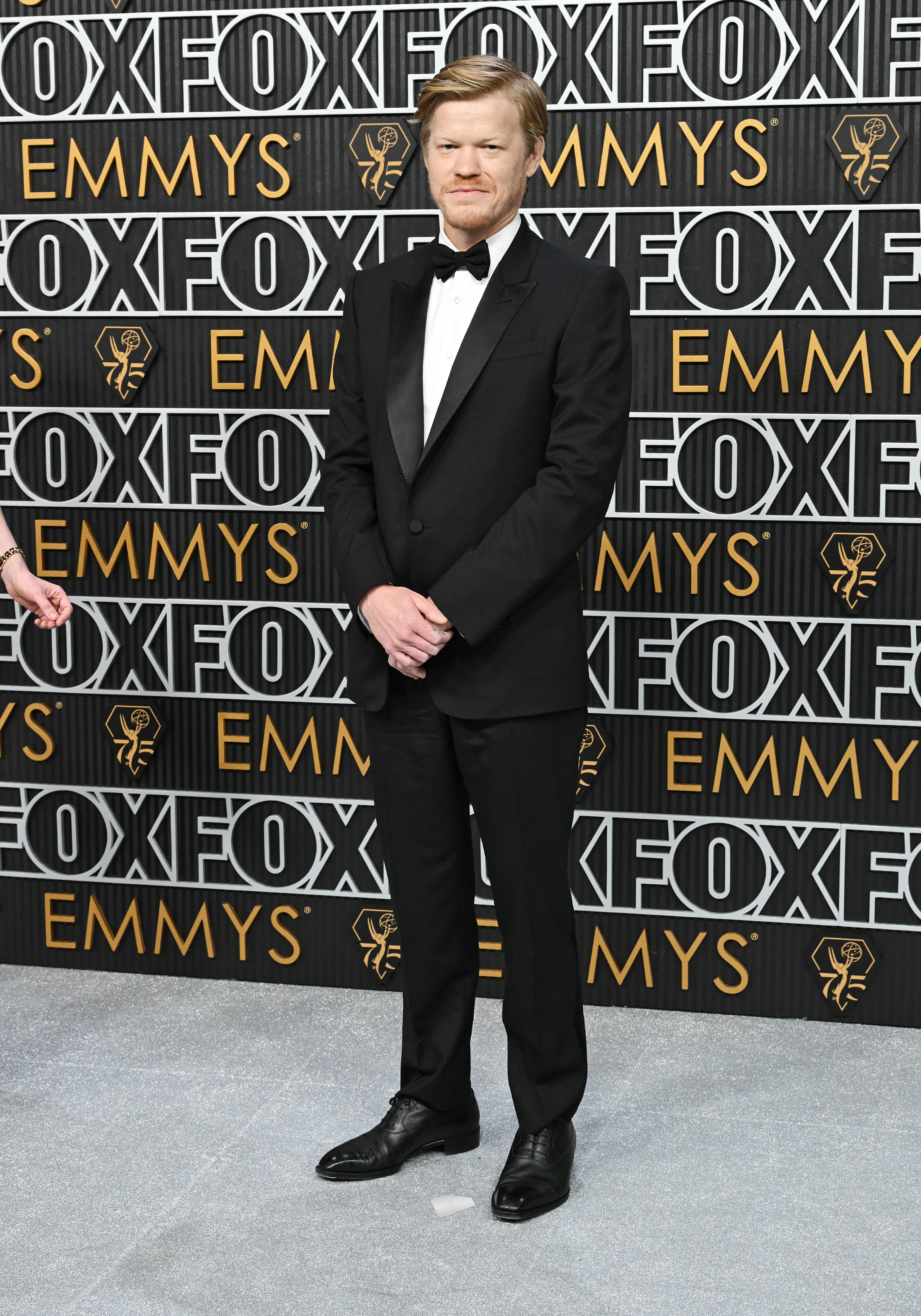 Jesse Plemons stands on the red carpet at the Emmys, wearing a classic black tuxedo with a bow tie. The backdrop features &quot;FOX&quot; and &quot;Emmys&quot; in repeated patterns