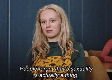 A person with blonde hair, wearing a green top and yellow pattern overalls, sits in a classroom with text overlay: &quot;People forget that bisexuality is actually a thing.&quot;