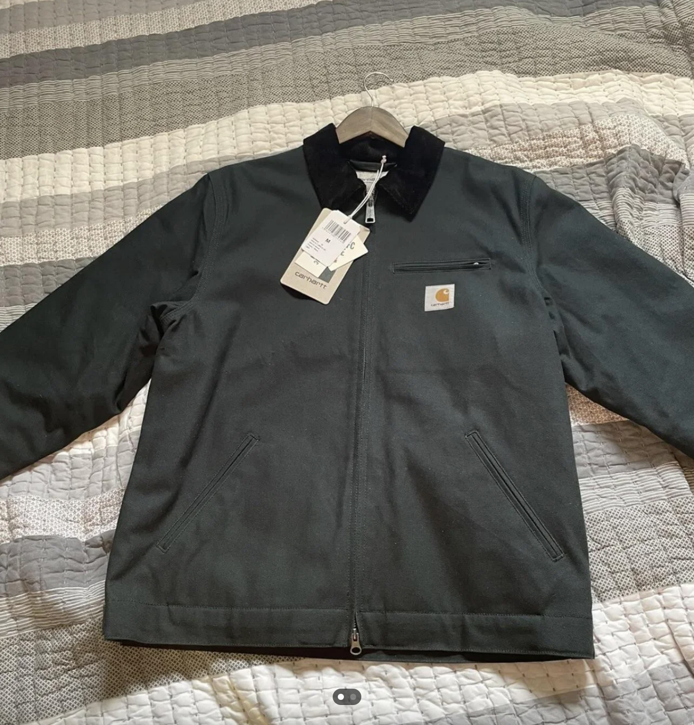 A dark jacket with tags attached, by Carhartt, displayed on a quilted bedspread