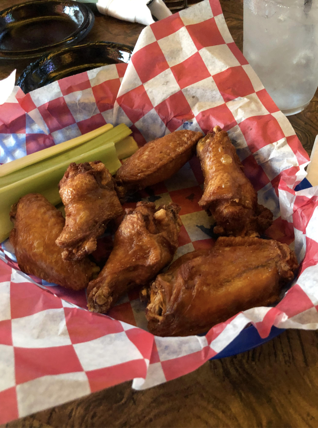 Basket containing chicken wings and celery sticks, lined with a red and white checkered paper, with a glass of water in the background
