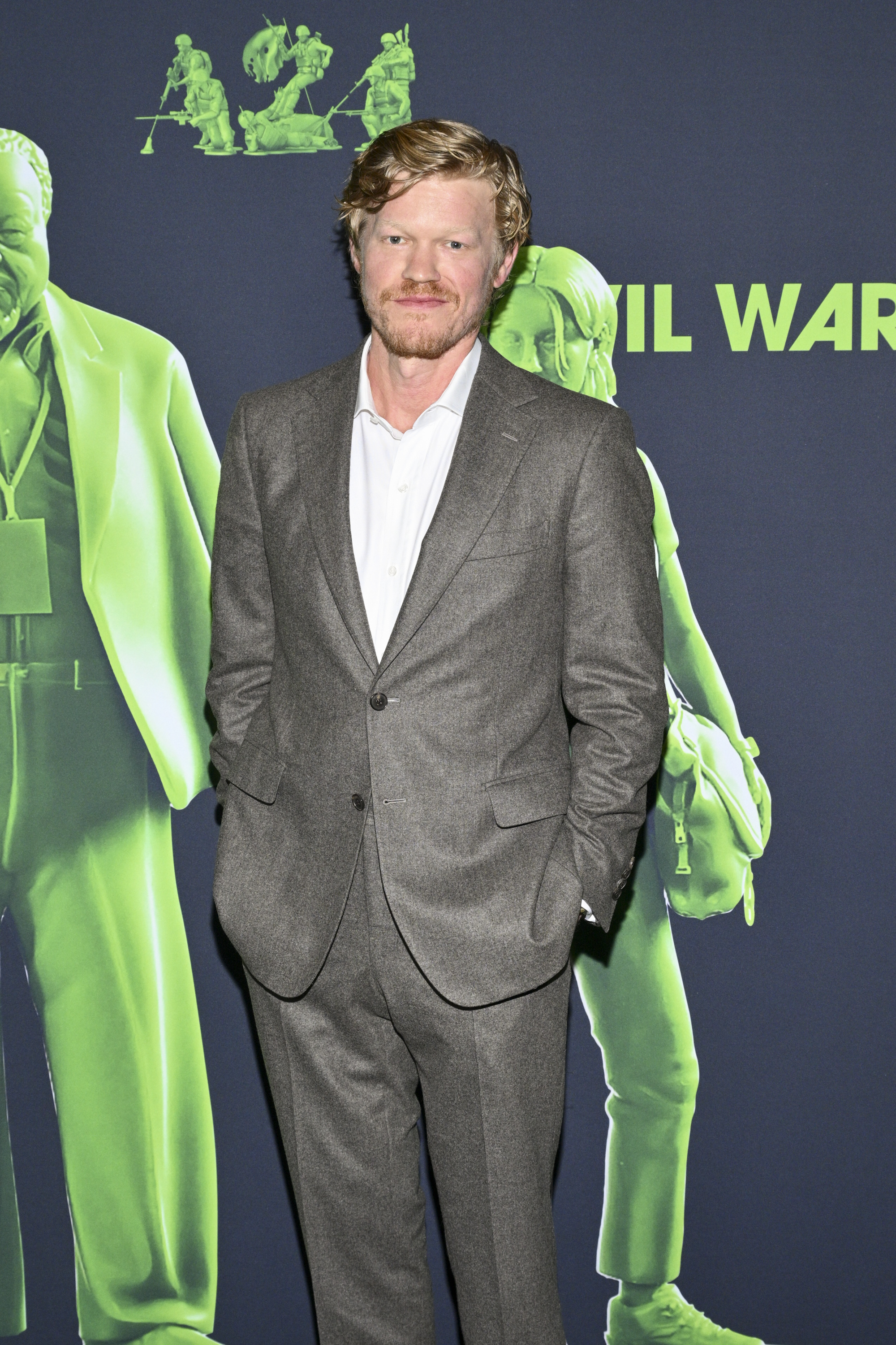 Jesse Plemons at an event, wearing a gray suit with a white shirt, standing in front of a backdrop featuring green figures and the text &quot;Civil War.&quot;