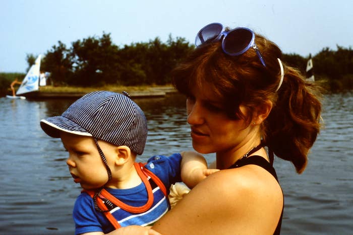 A mother with sunglasses on her head lovingly holds a baby wearing a striped cap near a lake with boats in the background. Their names are not known