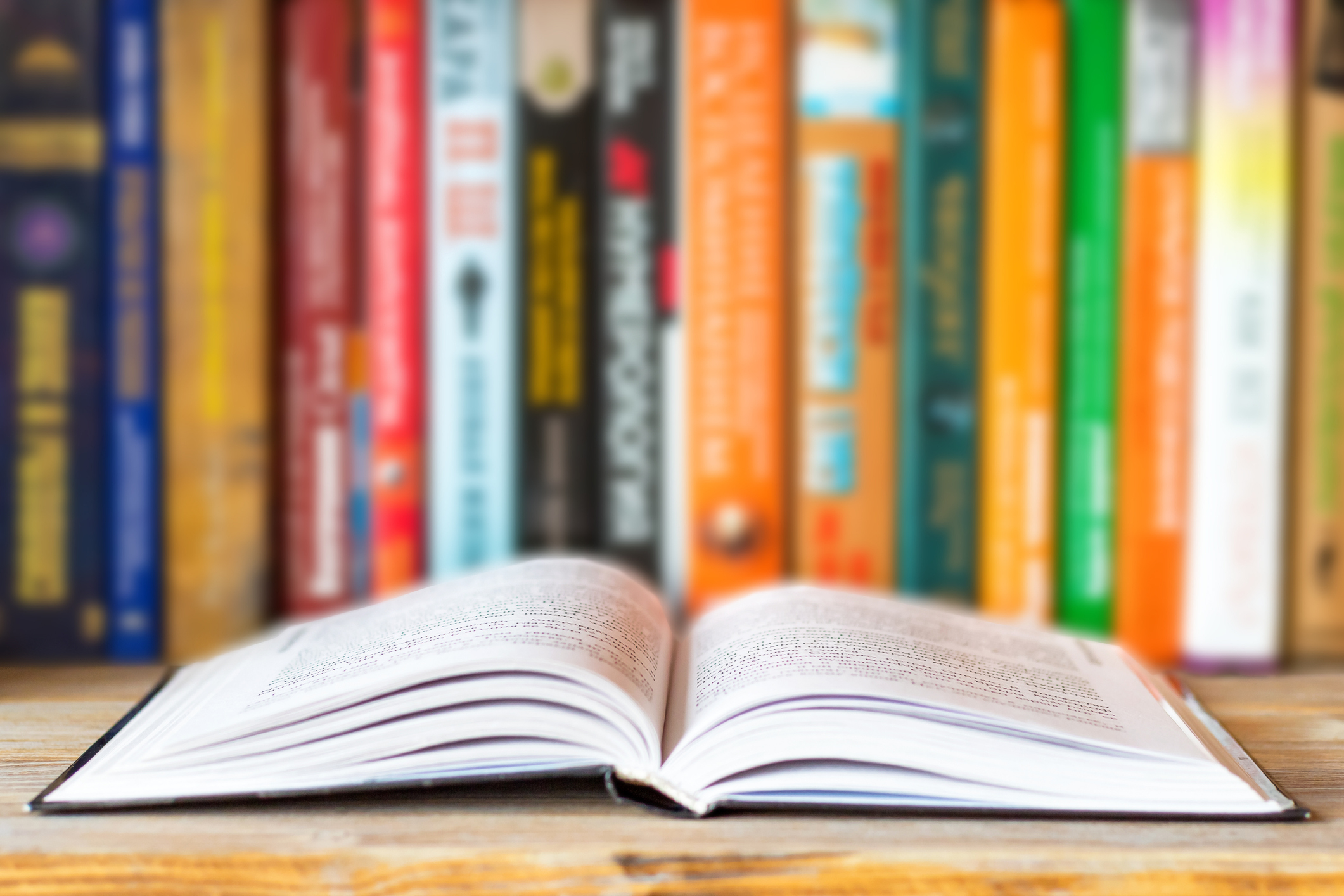Open book on a wooden surface with a background of colorful, blurred books on a shelf