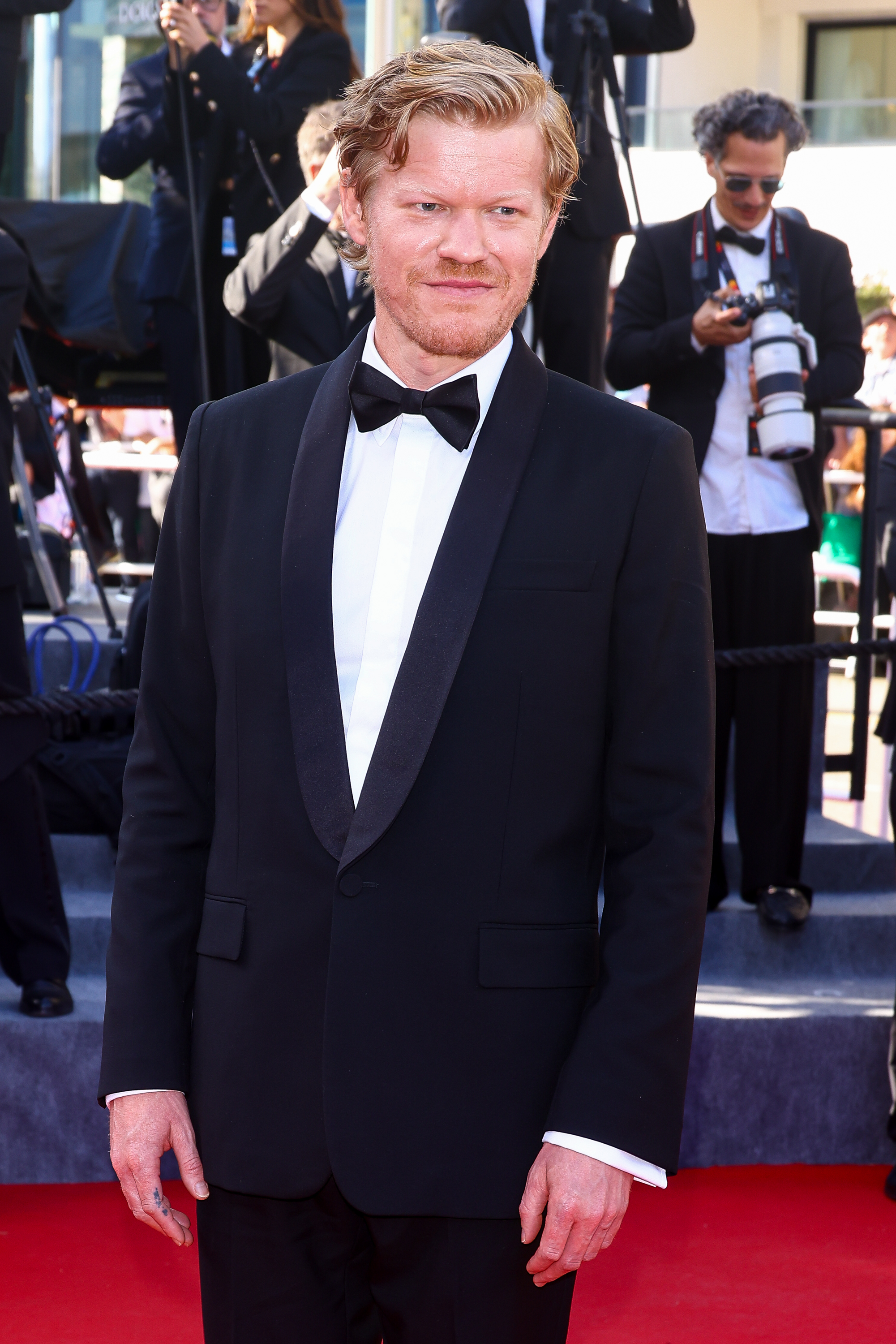 Jesse Plemons wearing a black tuxedo with a black bow tie, smiling on a red carpet with photographers in the background
