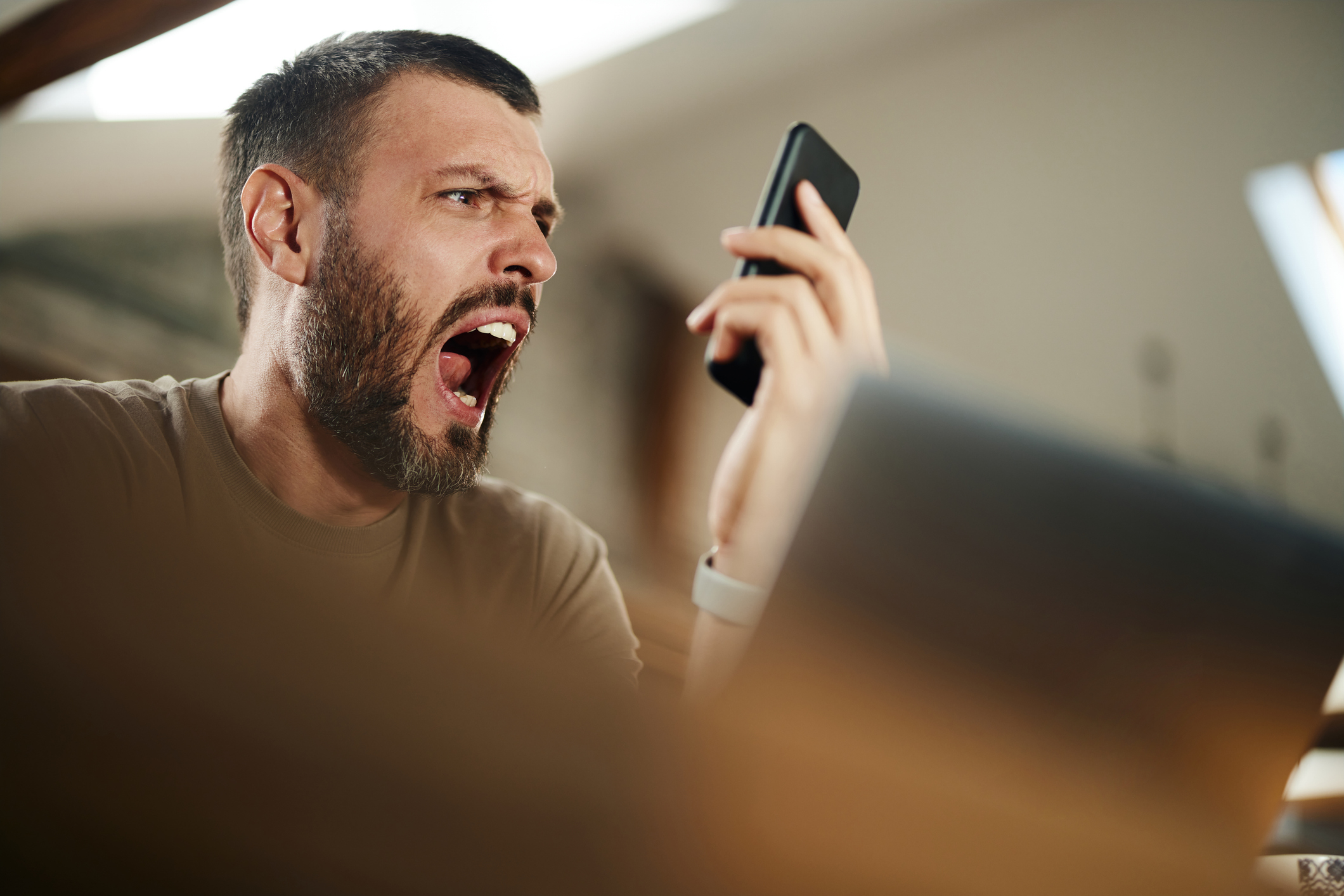 A man with a beard angrily yells into his smartphone while sitting indoors