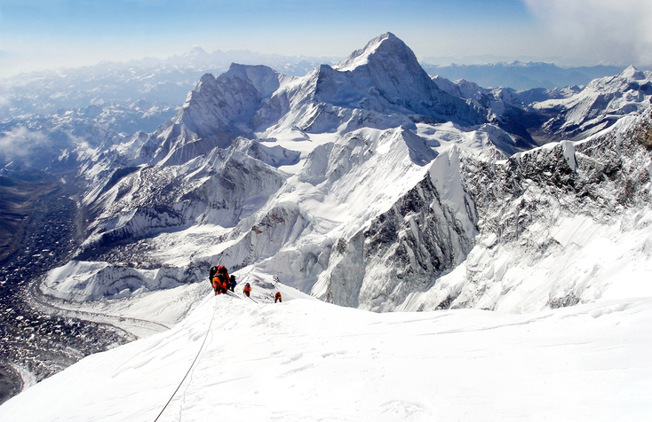 Mount Everest climbers ascend a snowy ridge with towering peaks in the background, connected by safety ropes amidst a vast mountain range