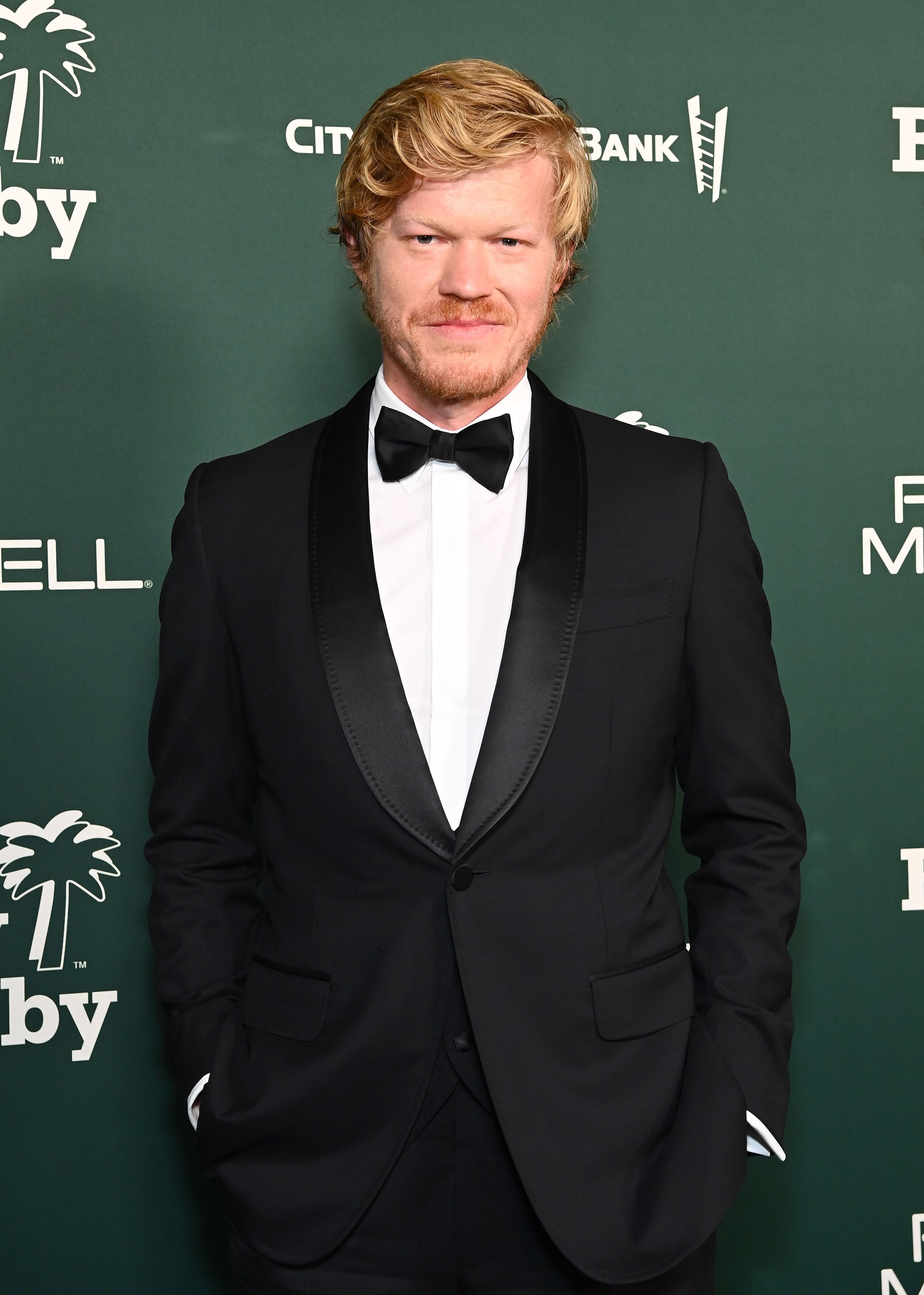 Jesse Plemons on a red carpet wearing a classic black tuxedo with a bow tie, standing against a green backdrop with sponsor logos