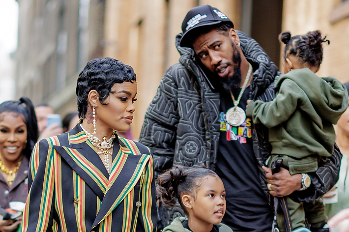 A group photo shows Teyana Taylor and Iman Shumpert with their children, as well as some friends, walking together. Teyana wears a striped blazer
