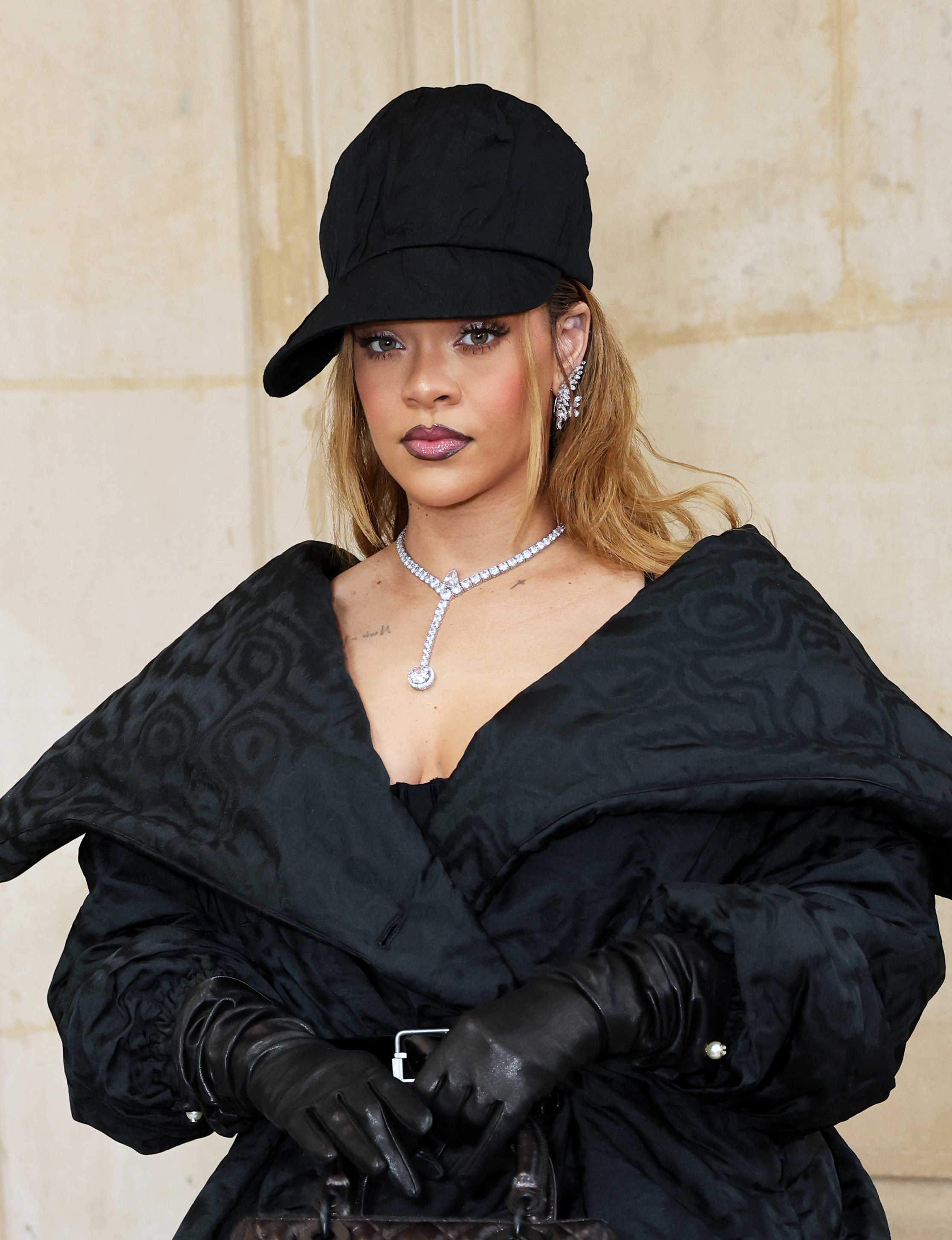 Rihanna is wearing a fashionable black ensemble, including a large coat, gloves, and a cap, accessorized with diamond jewelry, posing in front of a stone wall