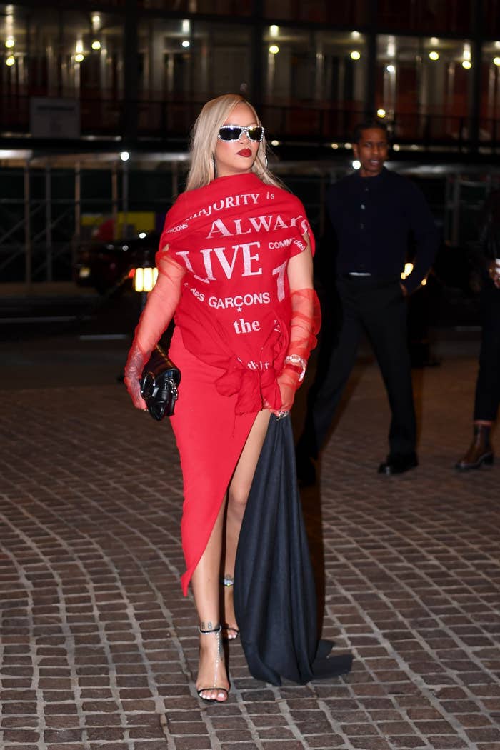 Rihanna in a bold, off-shoulder red dress with white text, holding a black clutch and wearing sunglasses. She walks confidently with a leg showing through a high slit