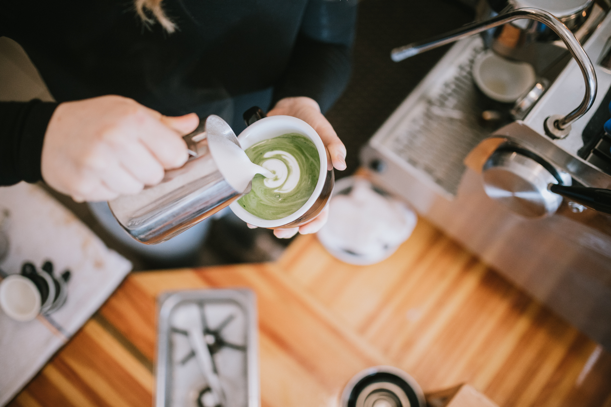 Barista pouring steamed milk into a cup of matcha tea, creating latte art at a coffee shop counter