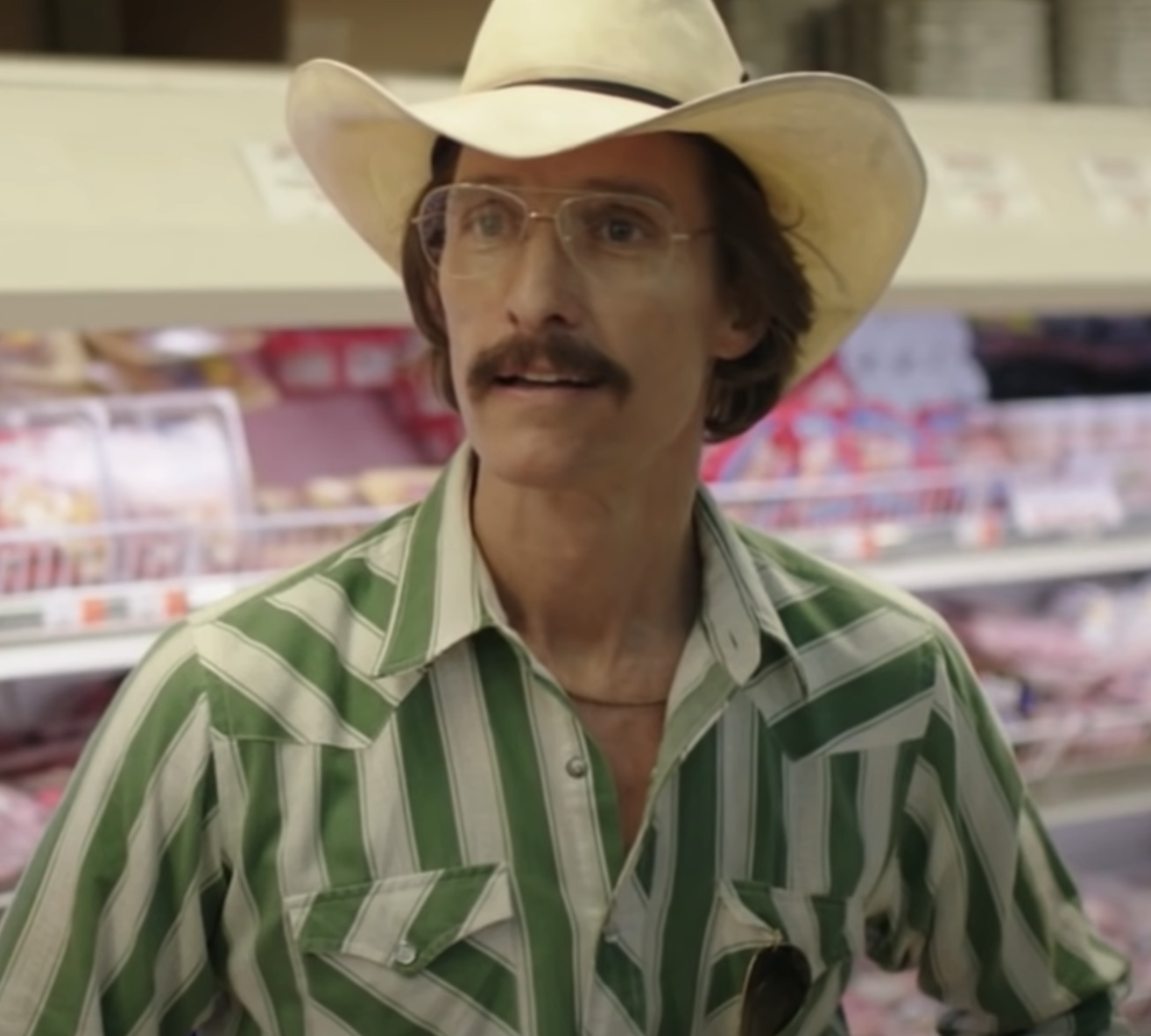 Matthew McConaughey in a grocery store, wearing a striped shirt, glasses, and a cowboy hat, standing in front of meat products