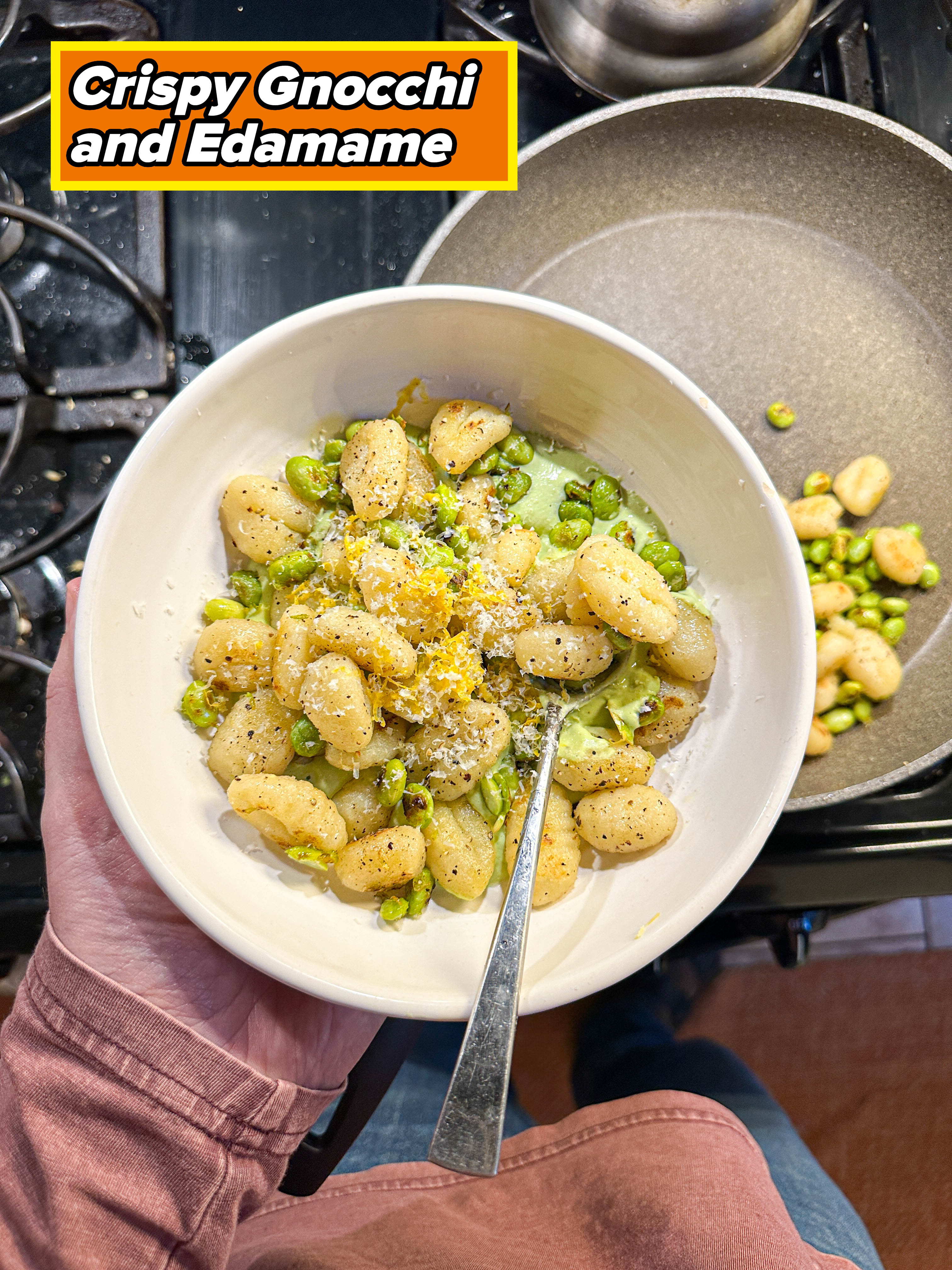 Hand holding a white bowl of gnocchi with peas and grated cheese over a stove with a frying pan nearby