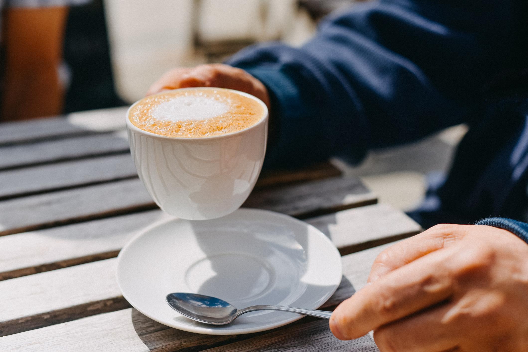 A person holding a cappuccino by the handle, with a saucer and spoon on a wooden table