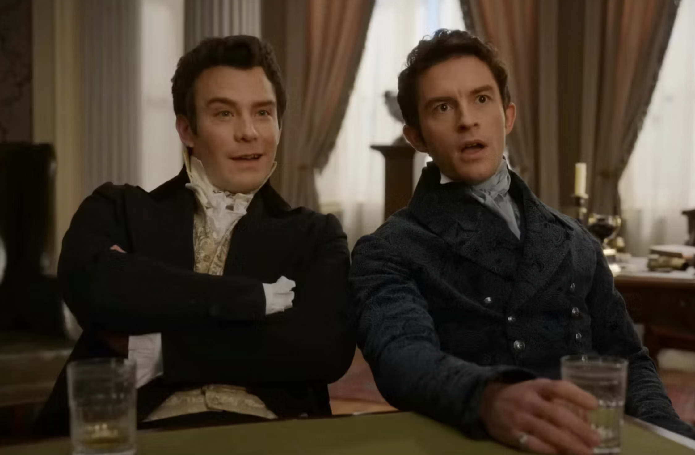 Jonathan Bailey y Luke Thompson, vestidos con trajes de época, están sentados y sosteniendo vasos en una escena de una serie de televisión