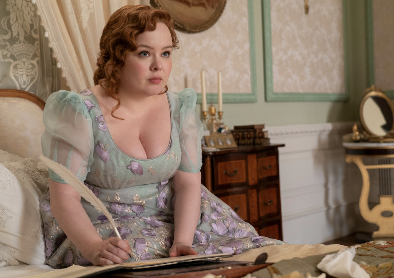 Nicola Coughlan, actriz de Bridgerton, en un vestido de época floral, escribiendo con una pluma en una habitación decorada al estilo antiguo