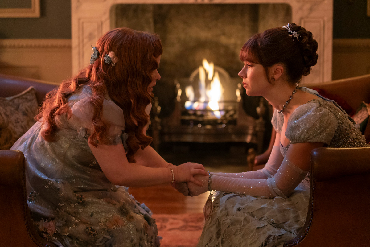 Nicola Coughlan y Claudia Jessie en una escena de "Bridgerton", vestidas con trajes de época mientras se sientan frente a una chimenea encendida