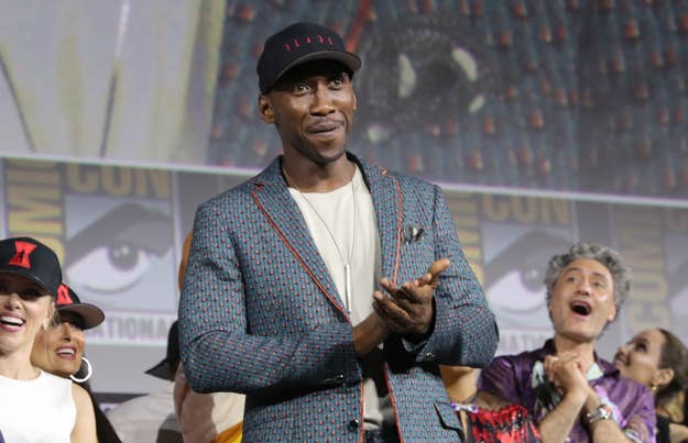 Mahershala Ali, in patterned suit and black cap, claps appreciatively on stage at a Comic-Con event with audience members and panelists looking excited