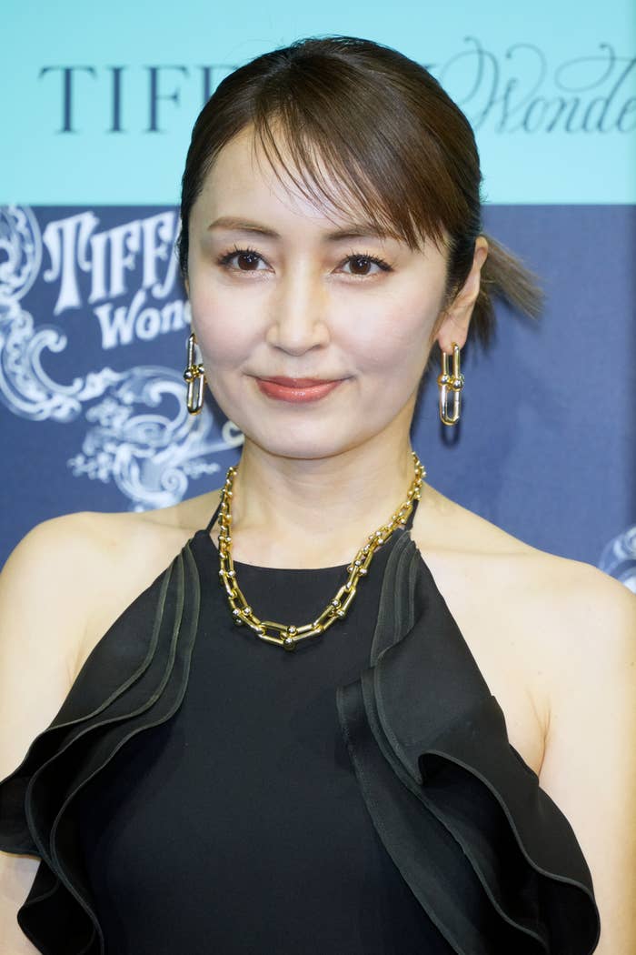 女性がカメラに向かって微笑んでいる。黒いドレスと金色のアクセサリーを着用している。名前は不明。