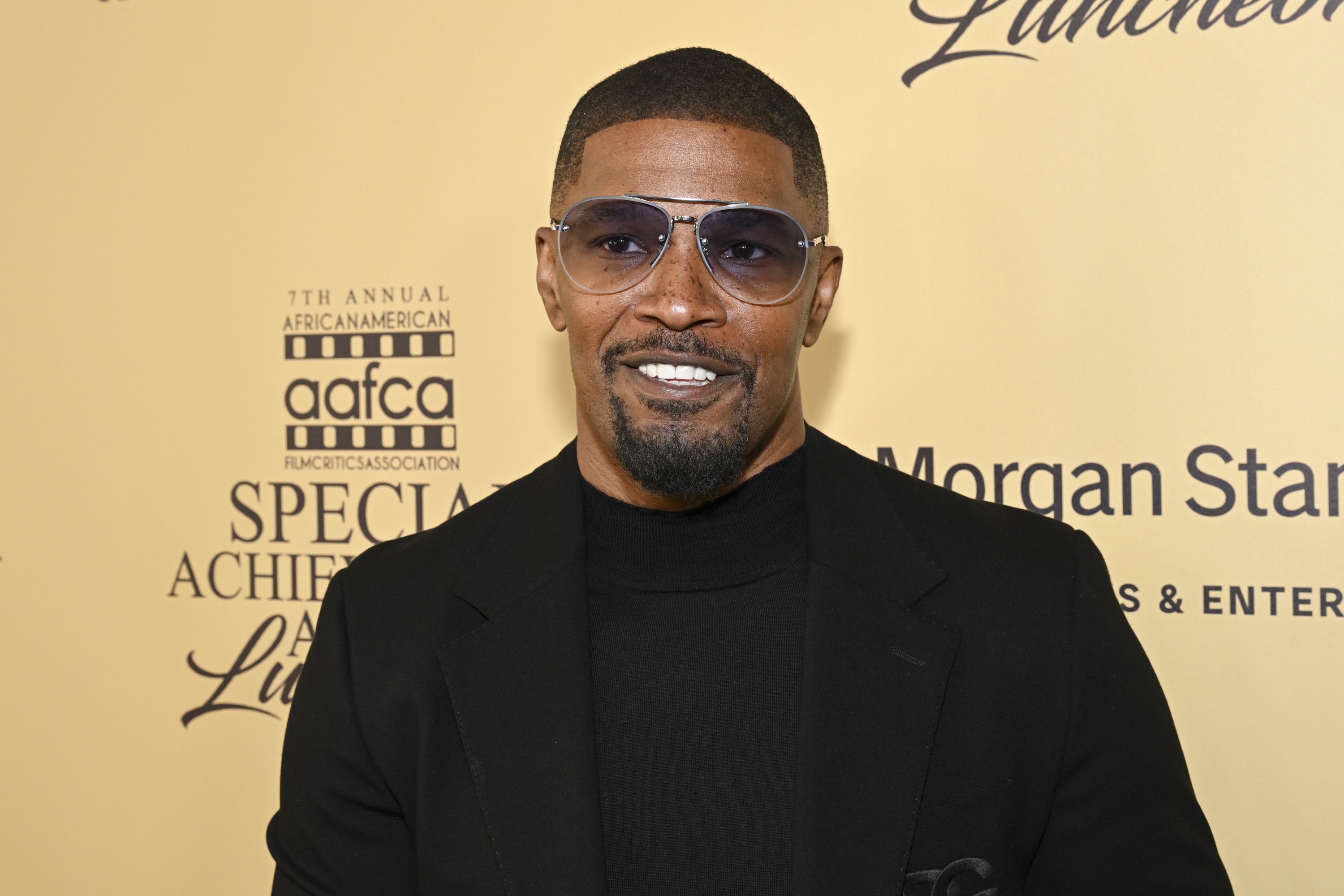 Jamie Foxx smiling, wearing a black turtleneck and blazer, standing in front of the backdrop for the 7th Annual African American Film Critics Association Special Achievement Luncheon