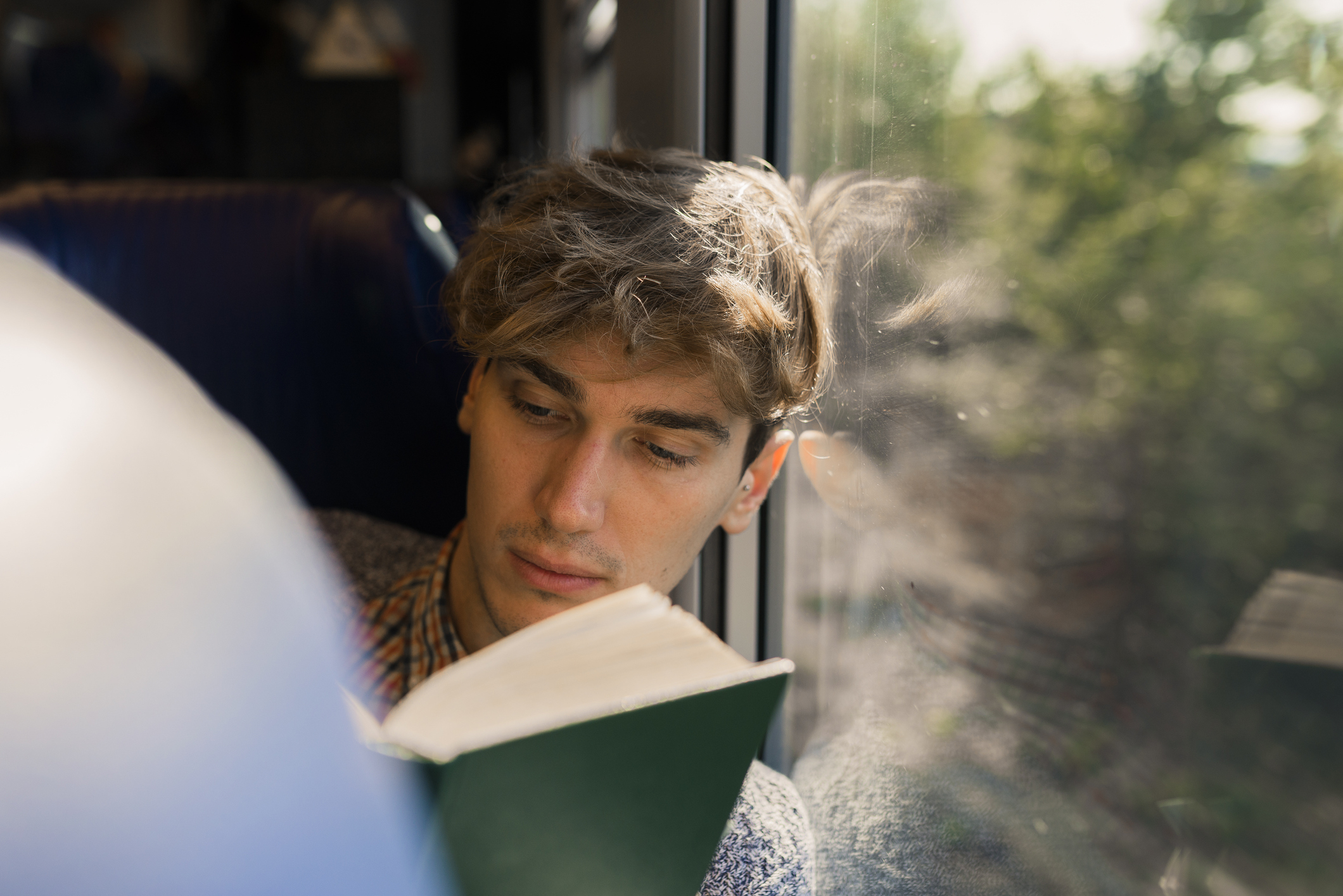 A person with wavy hair and a plaid shirt reads a book while sitting by a train window. Trees are reflected in the window outside.