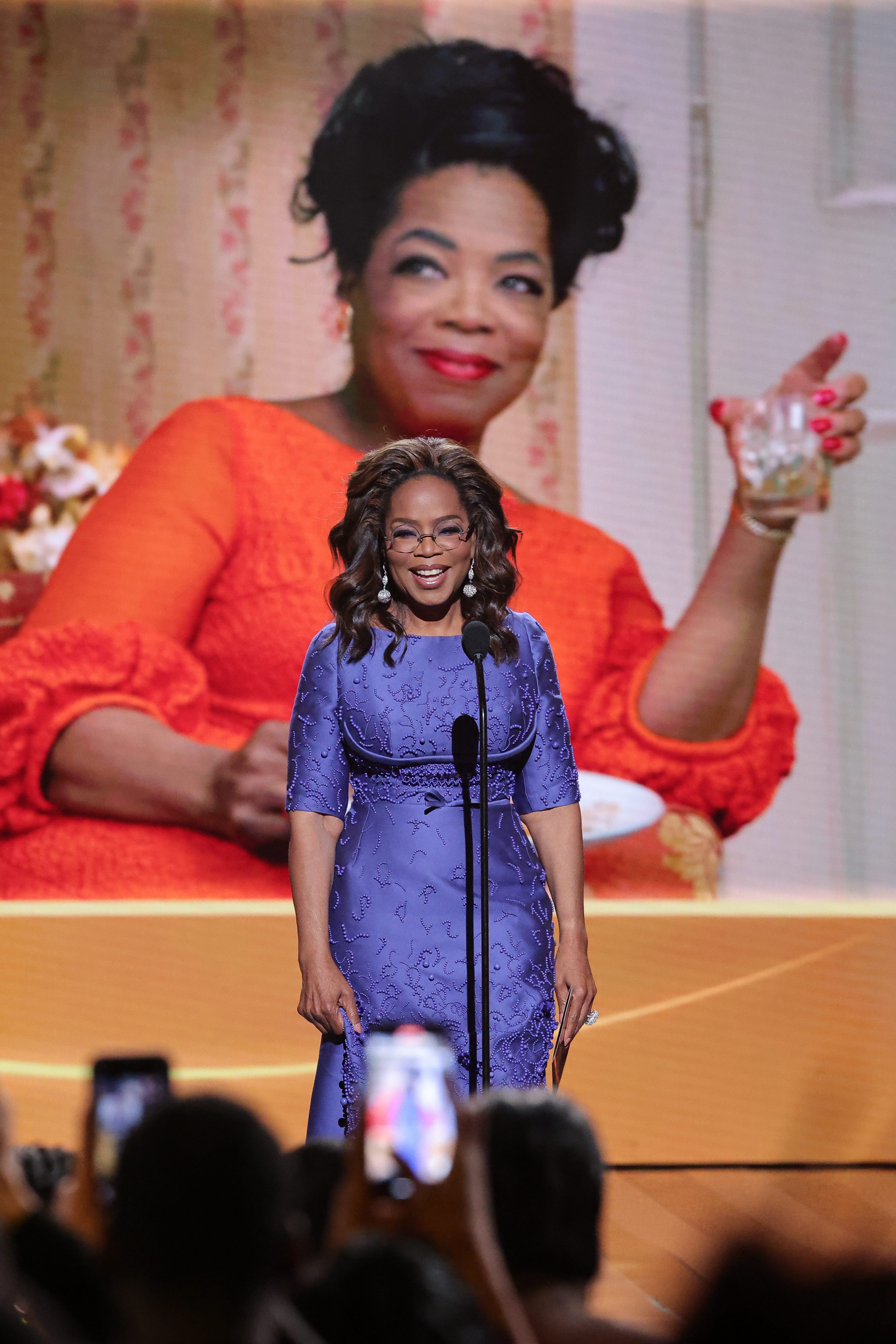 Oprah Winfrey stands on stage in a blue dress holding a microphone with a large image of herself in an orange outfit in the background