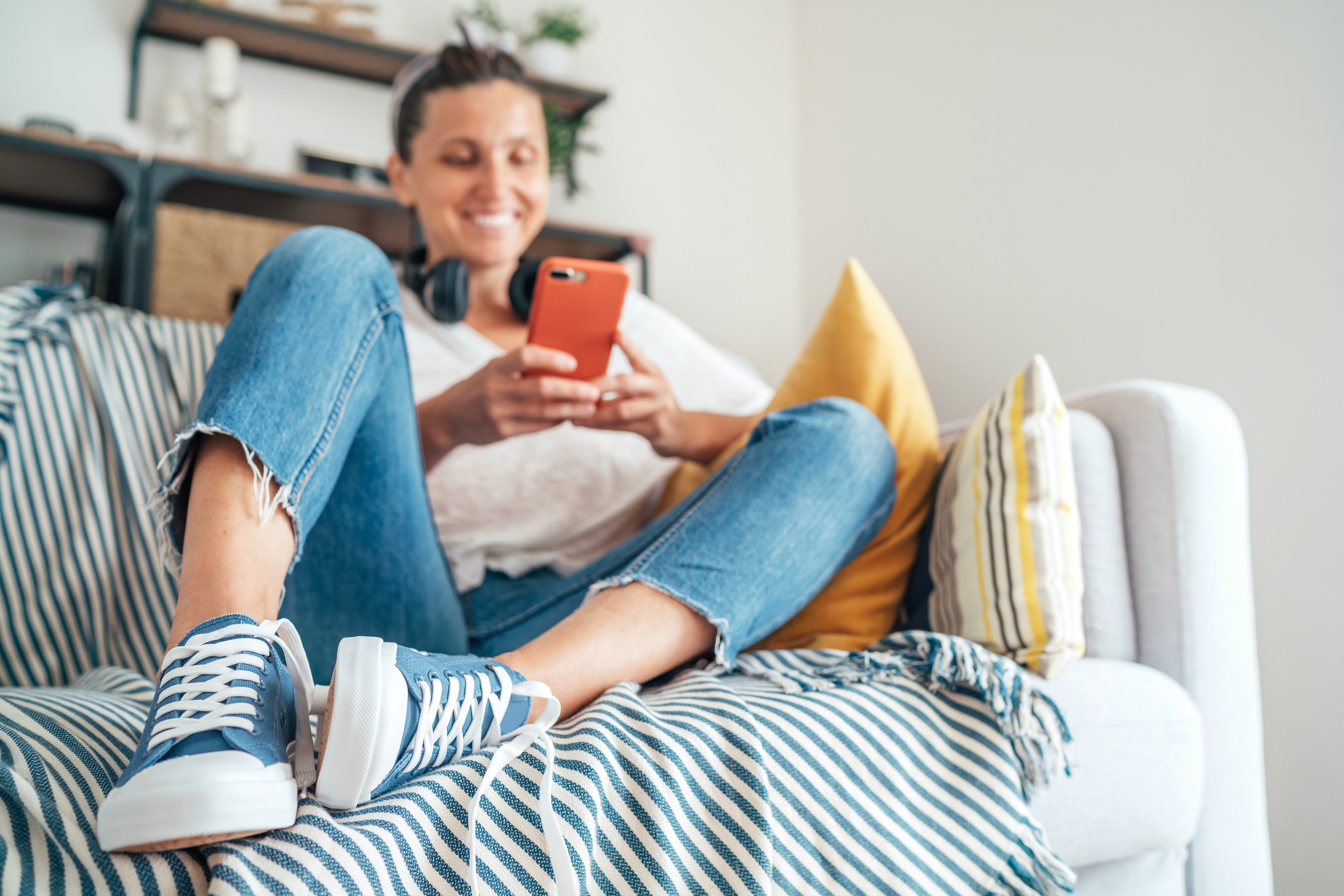 A woman is sitting comfortably on a couch, wearing ripped jeans, sneakers, a white top, and headphones around her neck, smiling while looking at her phone
