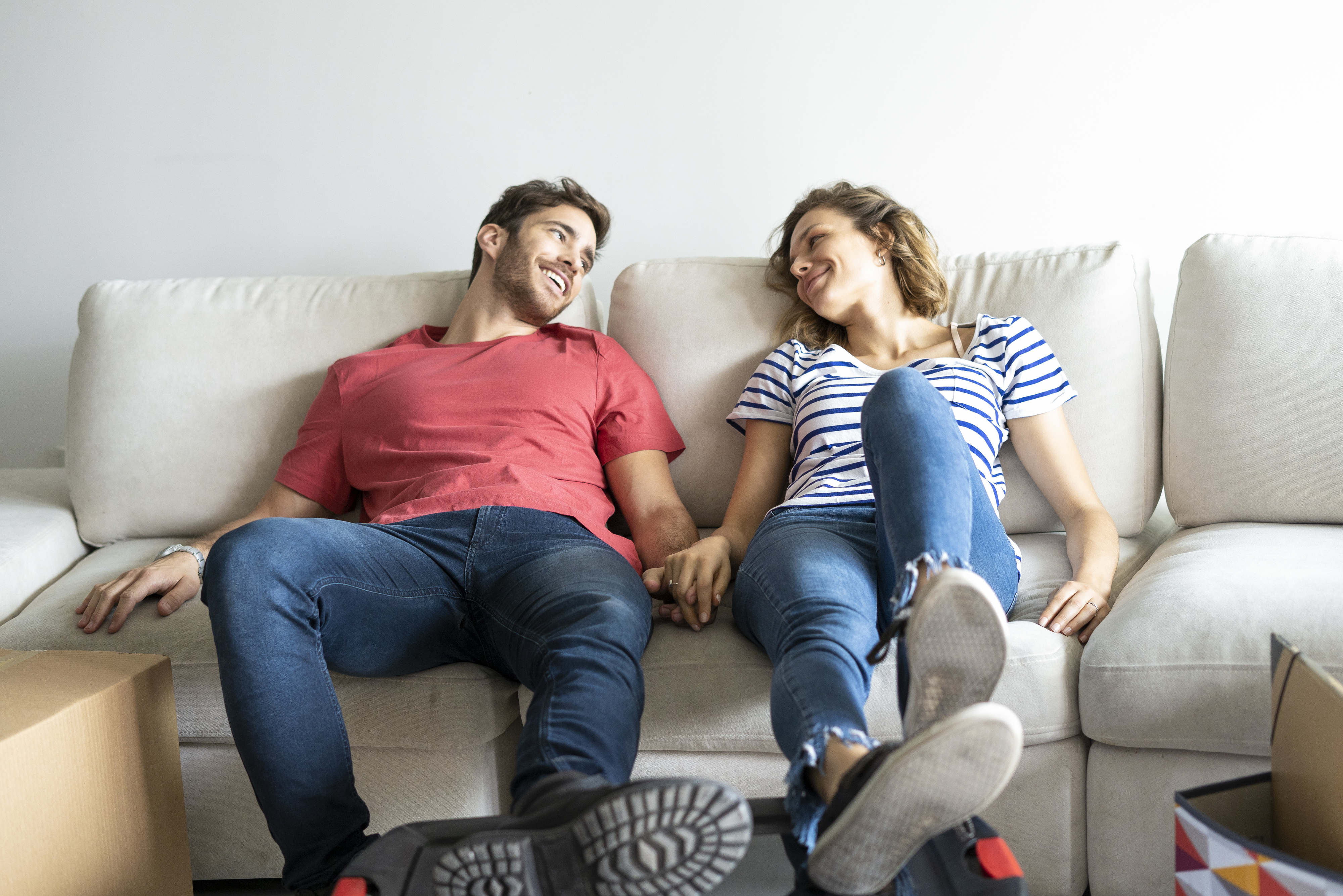 A couple, holding hands and smiling at each other, relaxes on a couch surrounded by cardboard boxes