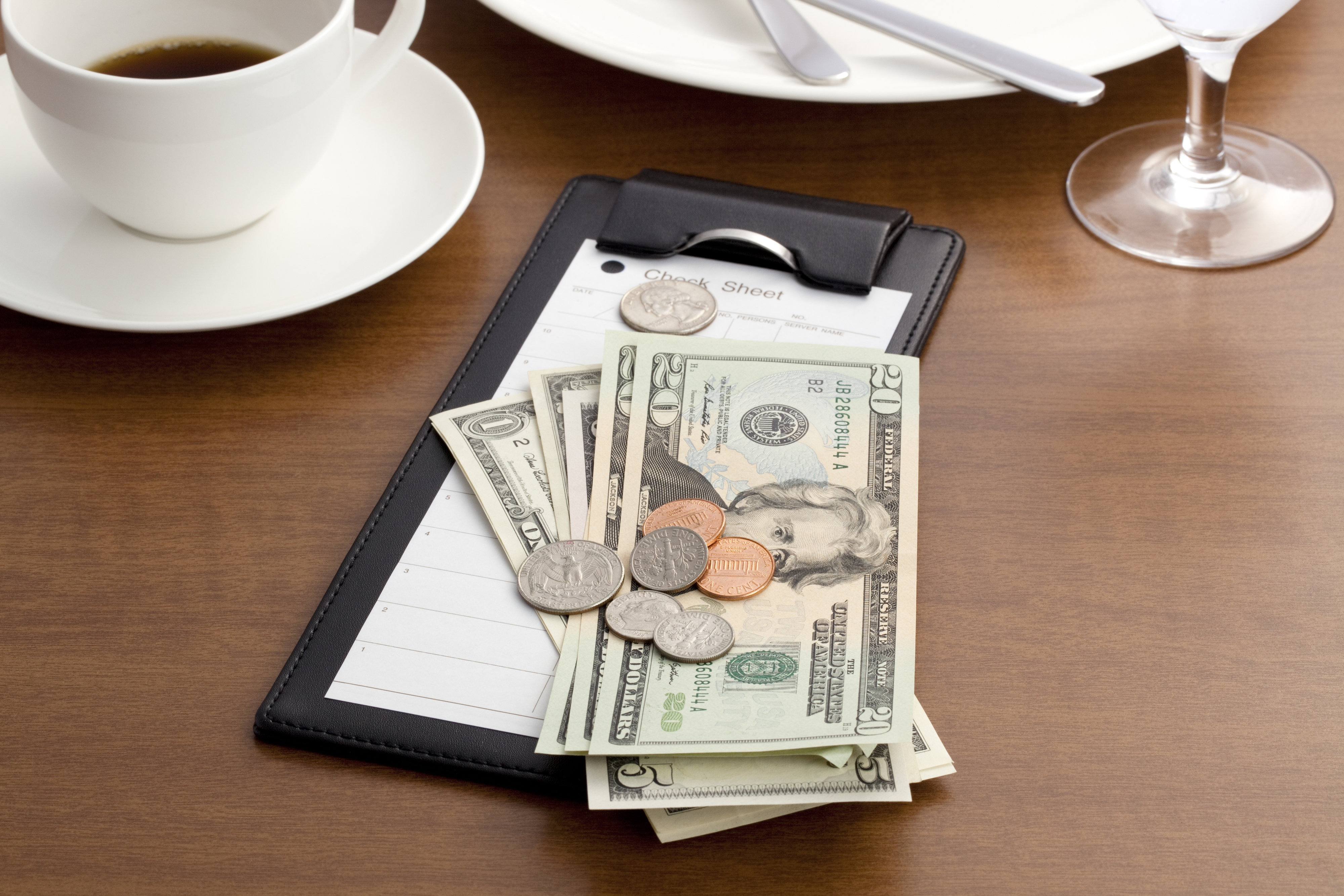A check sheet, various U.S. dollar bills, and coins are placed on a black check holder on a table next to a cup of coffee and a wine glass