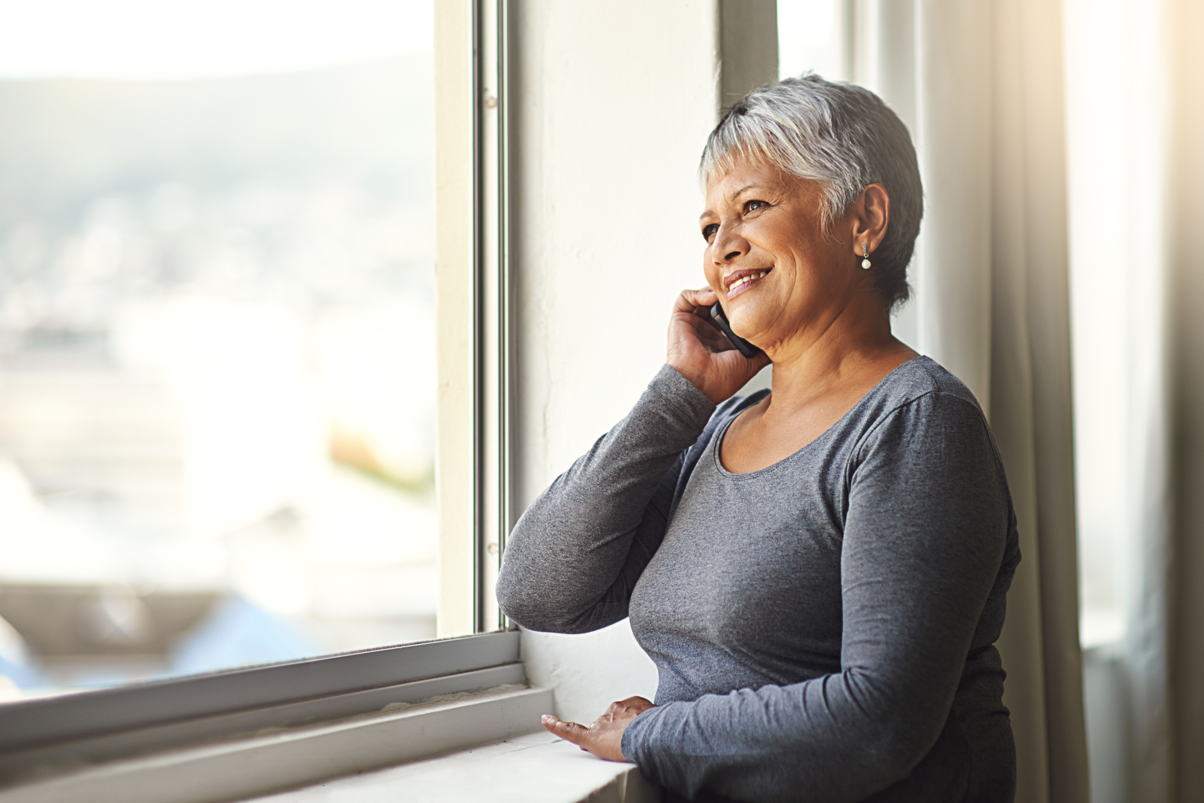 An older woman with short gray hair smiles while talking on a phone, standing by a window