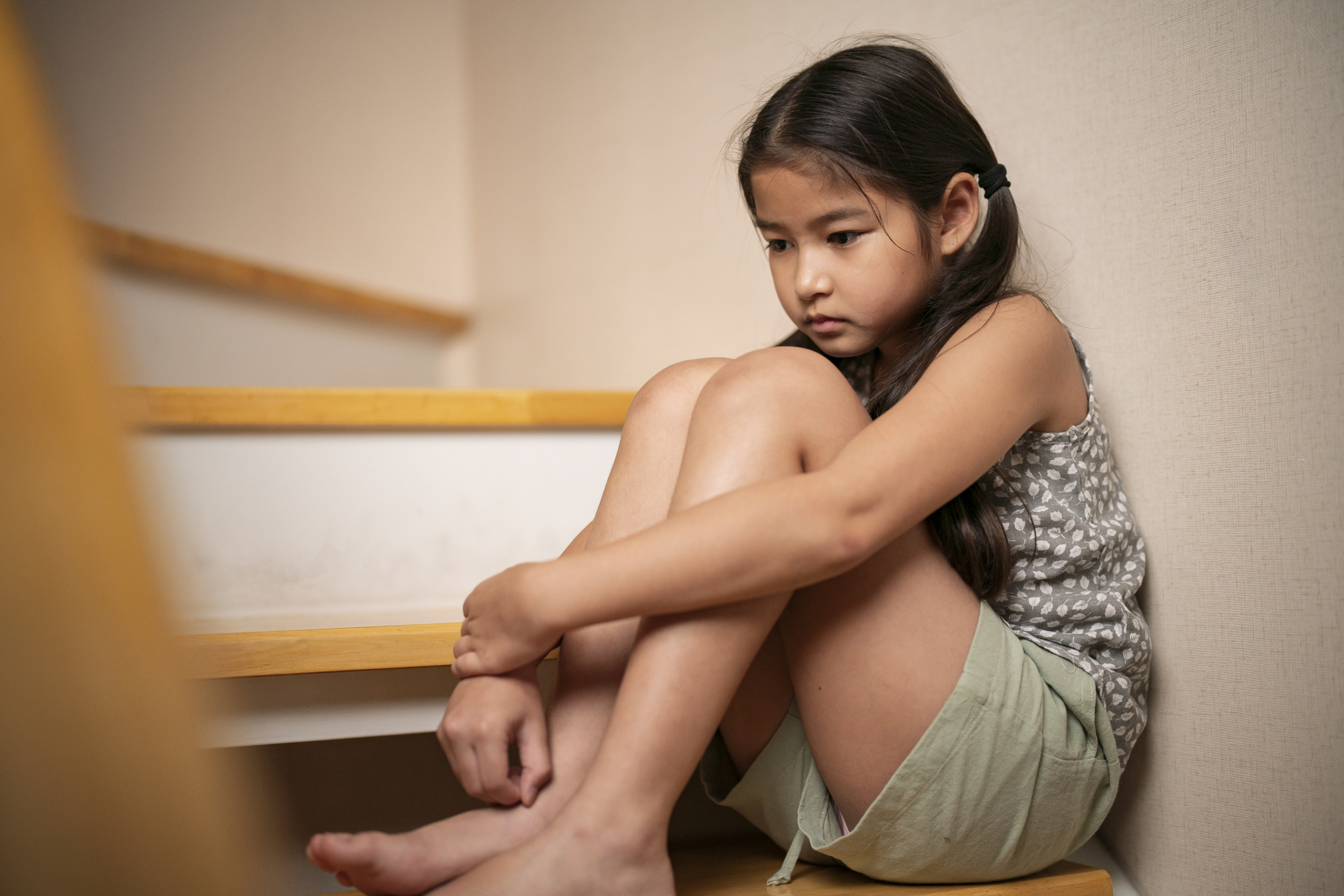 A young girl with long hair sits curled up on stairs, appearing sad. She is wearing a patterned tank top and shorts