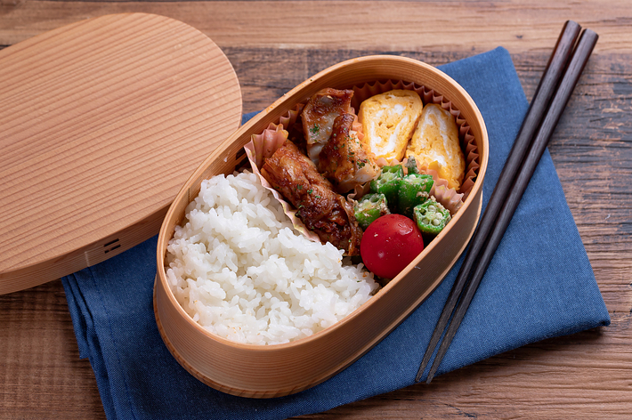 弁当箱にご飯、卵焼き、トマト、オクラ、そして肉料理が入っています。隣に箸があります。