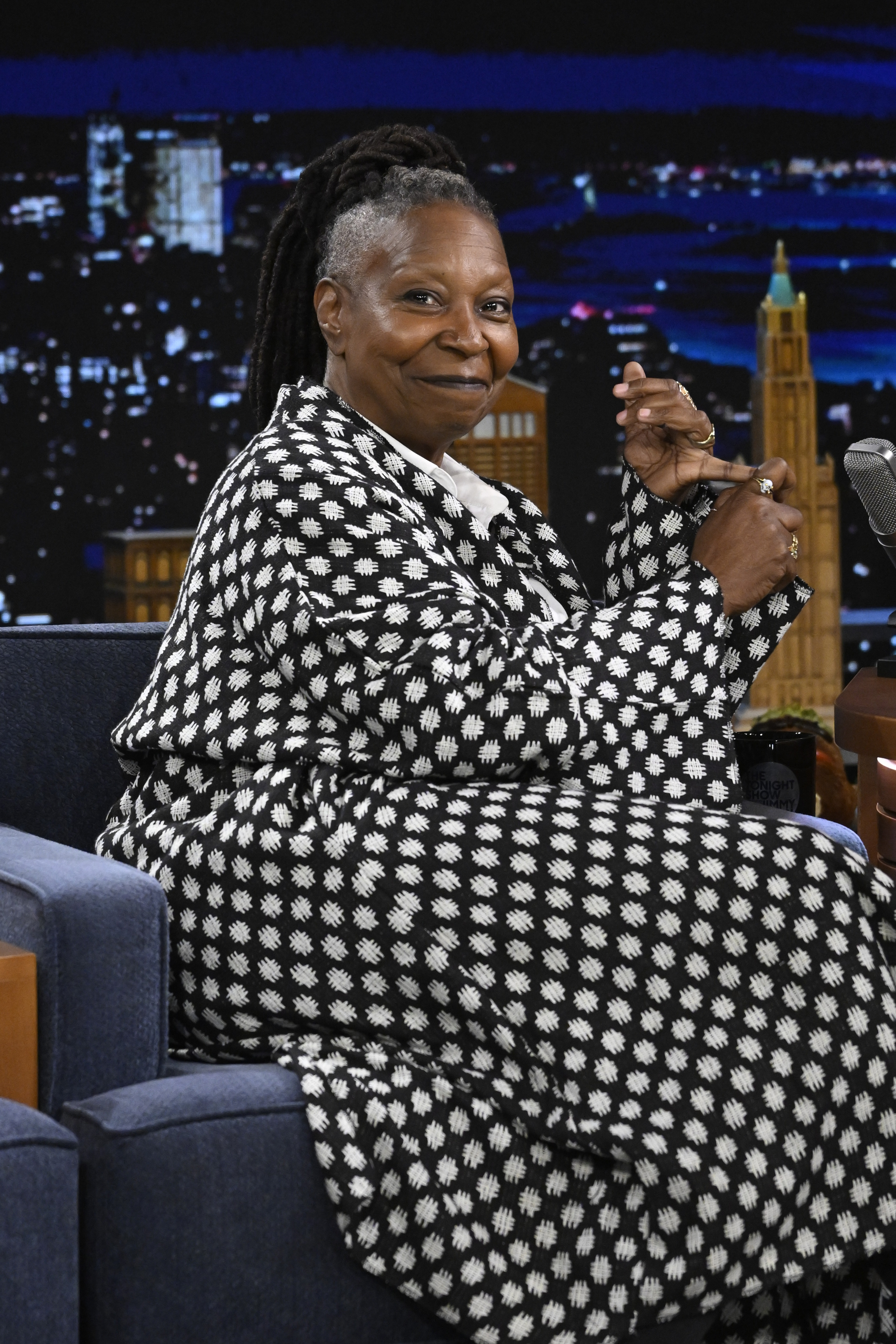 Whoopi Goldberg on a talk show set, wearing a patterned coat, sitting on a couch and smiling at the camera
