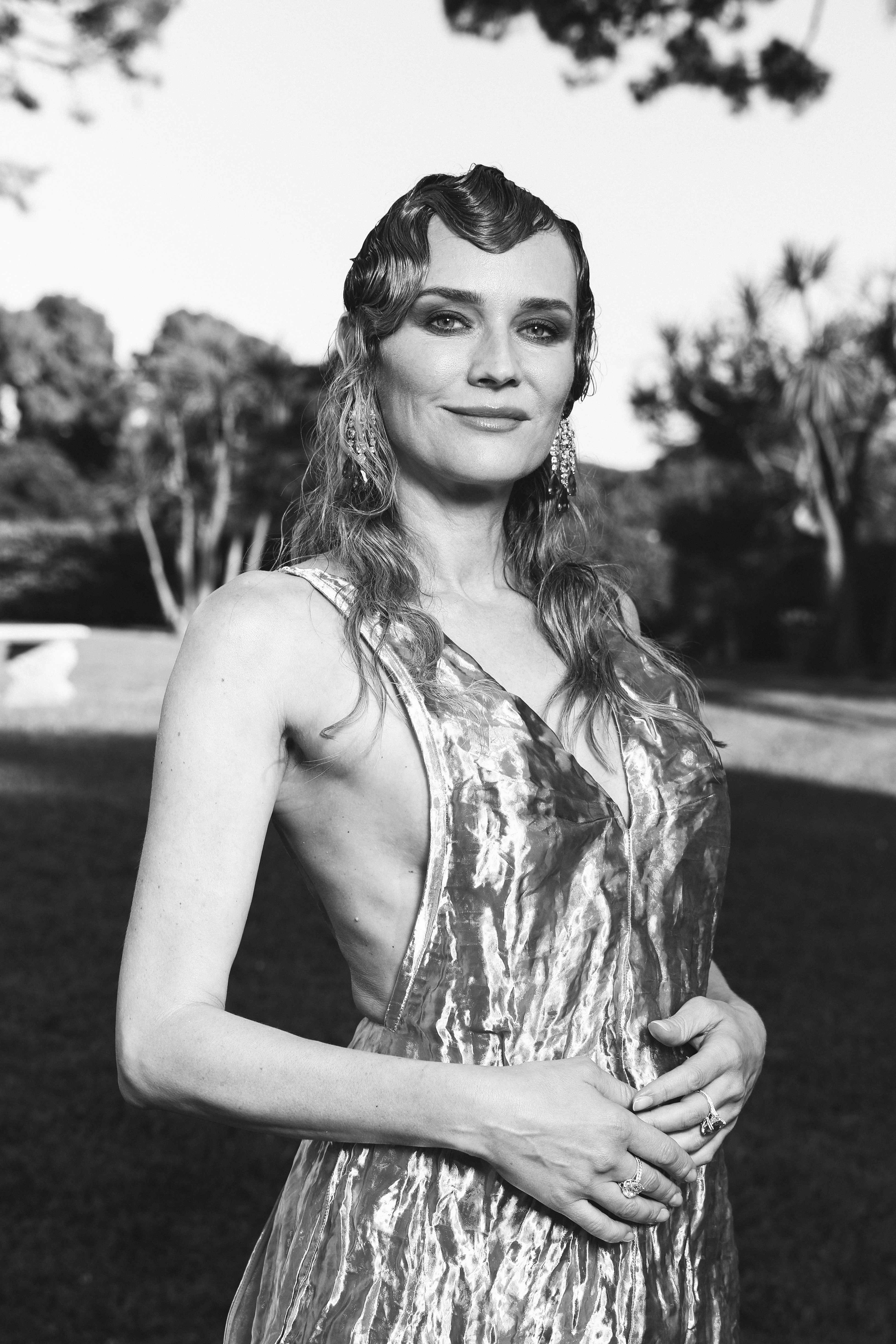 Diane Kruger wearing a sleeveless, textured dress, stands outdoors with hands clasped. Her hair is styled in waves and she wears large earrings