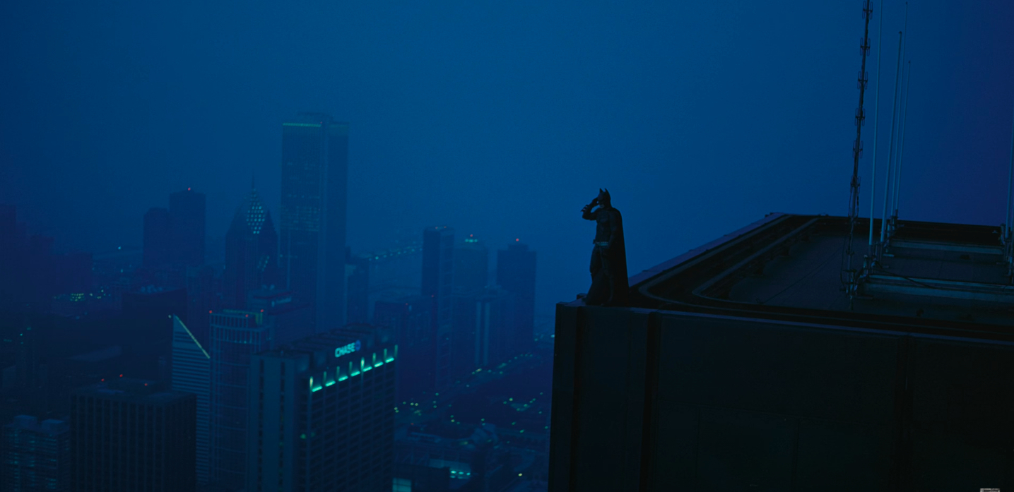 Batman stands on the edge of a tall building, overlooking a cityscape enveloped in dark blue mist