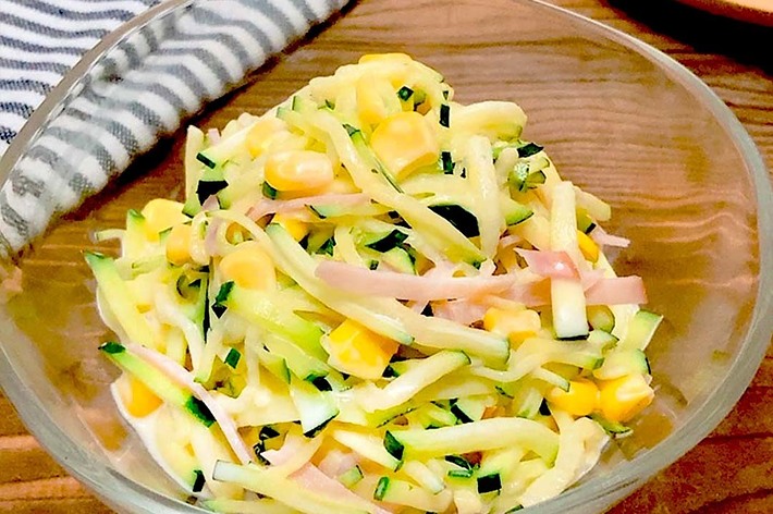 透明のボウルに盛られた、きゅうりとコーン、ハムのサラダ。横にはストライプの布が敷かれている。