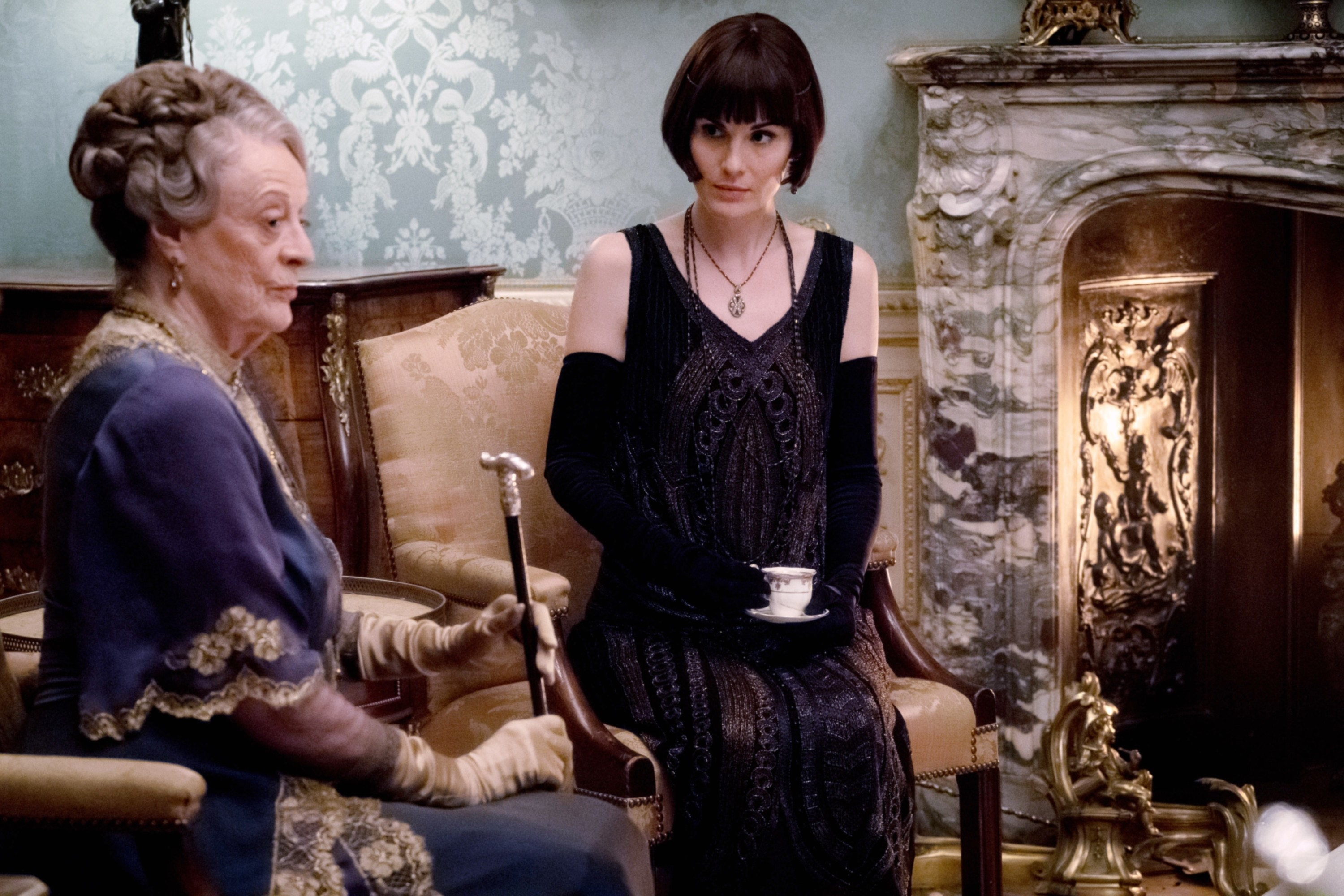 Maggie Smith and Michelle Dockery sit in an ornate room with period decor. Smith holds a cane, and Dockery wears an elegant black gown with gloves, holding a teacup