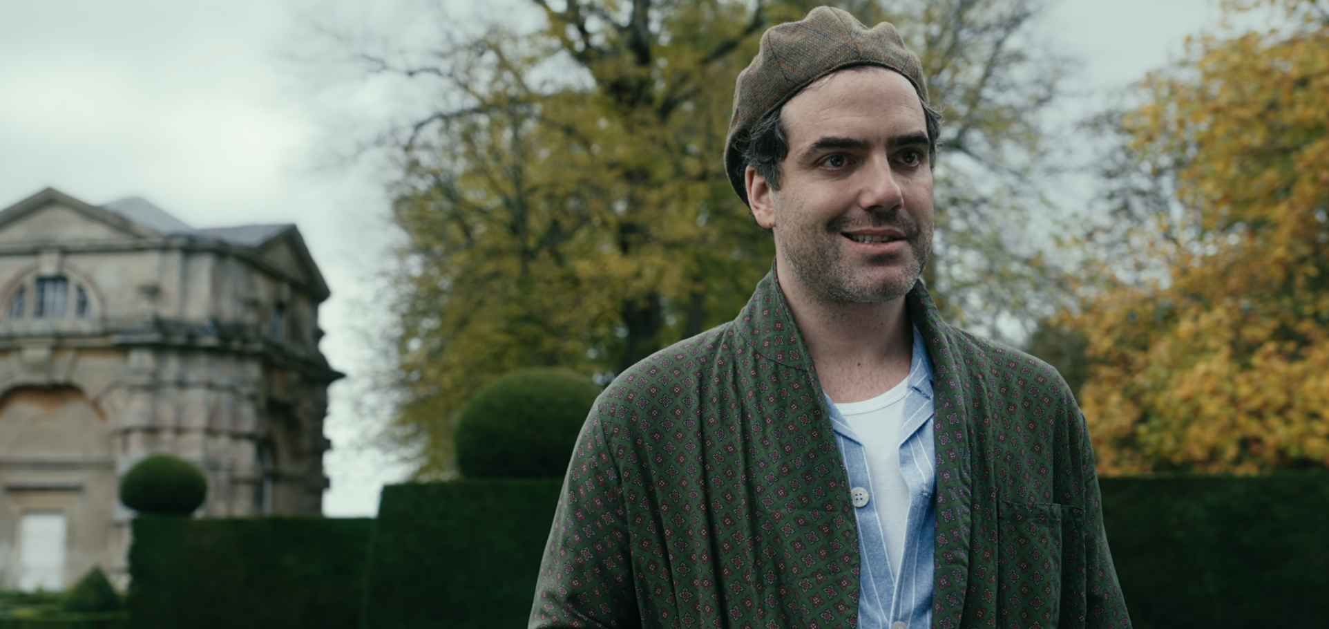 Man standing outdoors in front of a historical building and trees, wearing a jacket, shirt, and cap, smiling at the camera