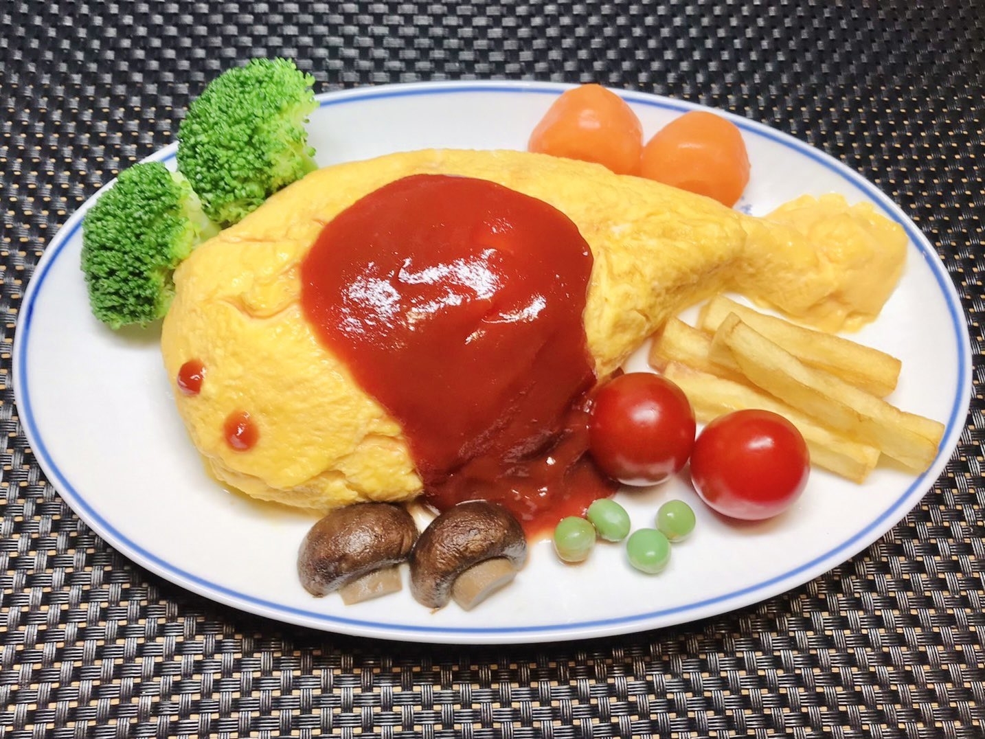 皿にはオムライスが盛られ、ケチャップソースがかけられています。添え物としてブロッコリー、ミニトマト、フライドポテト、マッシュルーム、グリーンピースが配置されています。