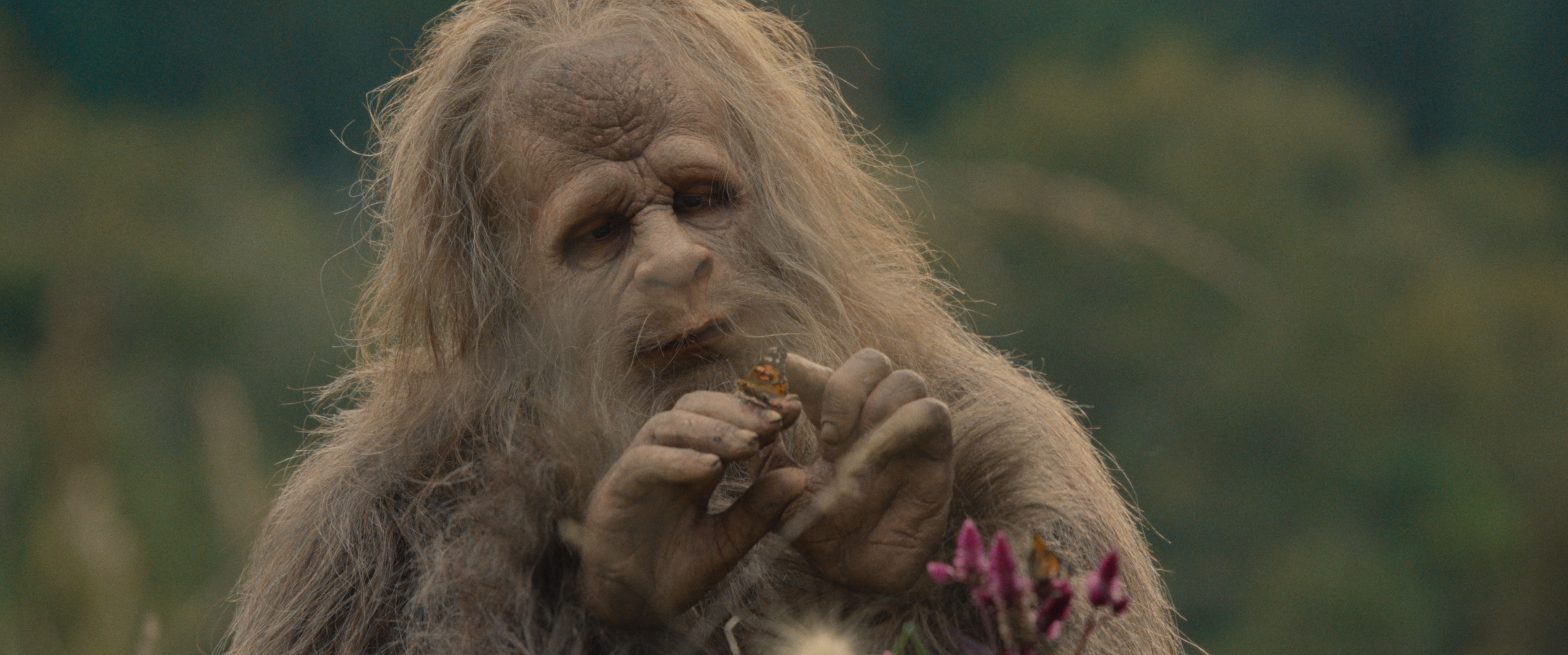 A creature resembling Bigfoot examines a flower outdoors, surrounded by greenery