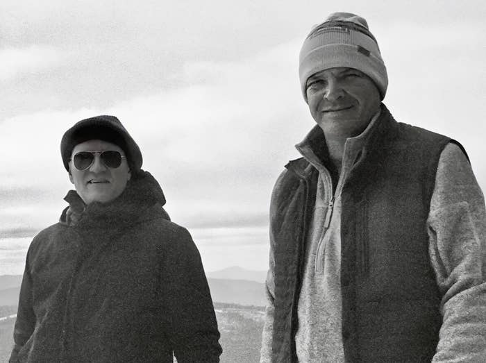 Two men, wearing winter clothes and hats, stand outside on a cloudy day. One man is smiling and the other wears sunglasses