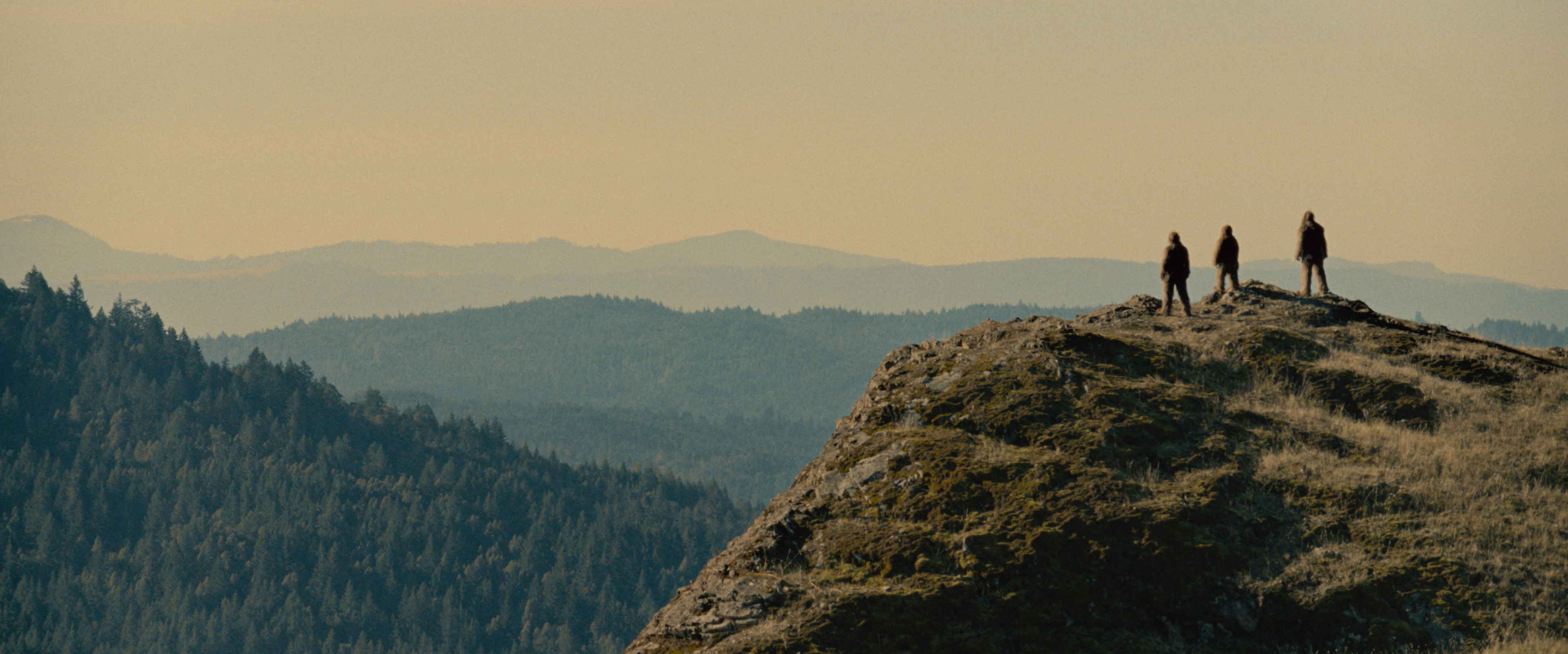 Three people stand on a rocky hilltop overlooking a vast landscape of forested mountains in the distance