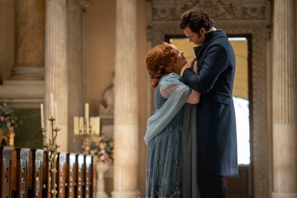 Nicola Coughlan and Luke Newton stand facing each other affectionately in an opulent room with tall columns, wearing historical period costumes