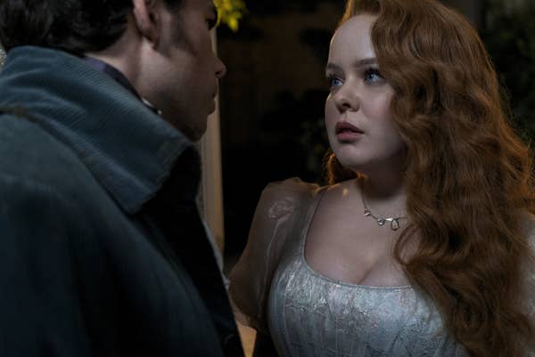 Nicola Coughlan and Luke Newton are gazing at each other, she is wearing a vintage-style dress and a necklace; he is in period costume