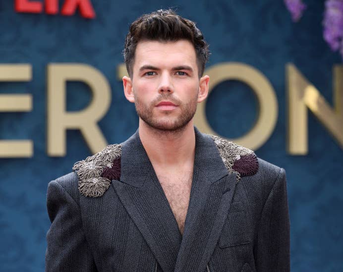 Luke Newton stands on the red carpet wearing a tailored, textured suit with intricate shoulder detailing