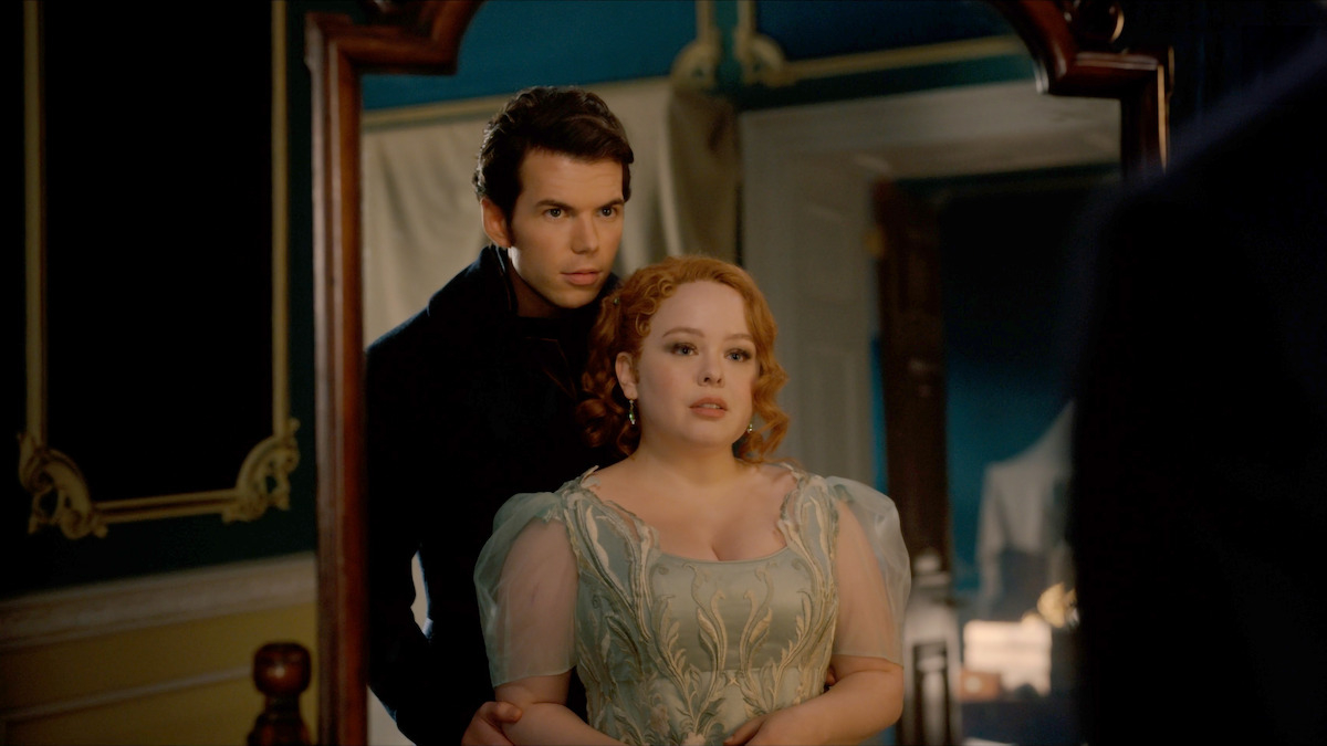 Luke Newton and Nicola Coughlan dressed in Regency-era attire, standing in front of a mirror, possibly a scene from a period drama