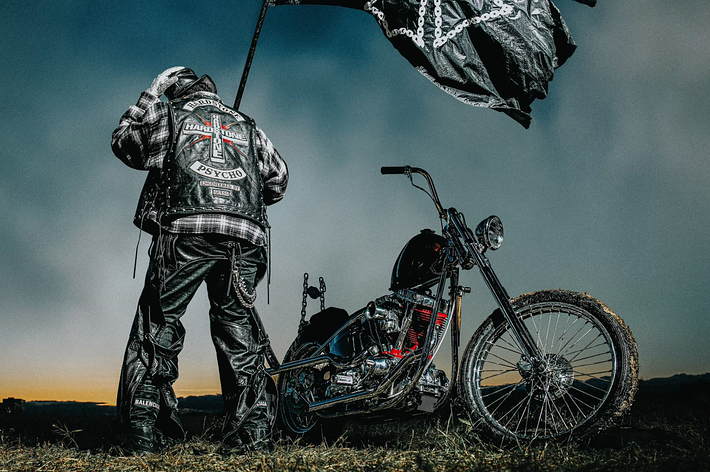 A biker in leather gear stands by a custom motorcycle, holding a flag. The back of the biker's jacket reads "Hardcore Carnivore Psycho." The image is related to music