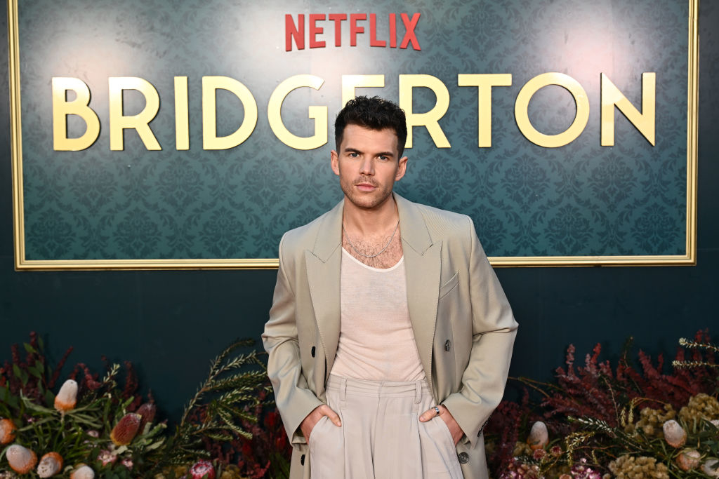 Luke Newton at the Bridgerton Netflix premiere, wearing a light-colored, tailored suit with a sheer top underneath