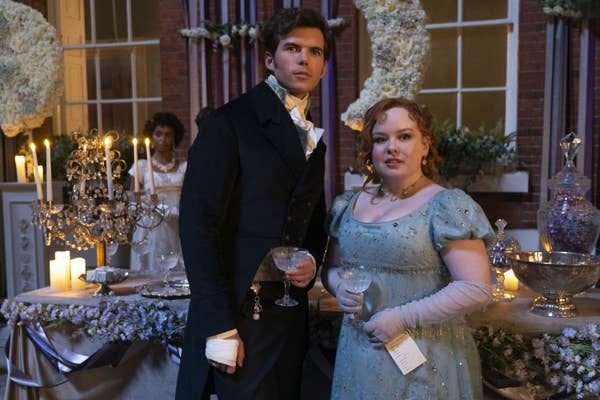 Luke Newton and Nicola Coughlan in period costumes, holding drinks at an elegant, candle-lit event with floral and antique decorations